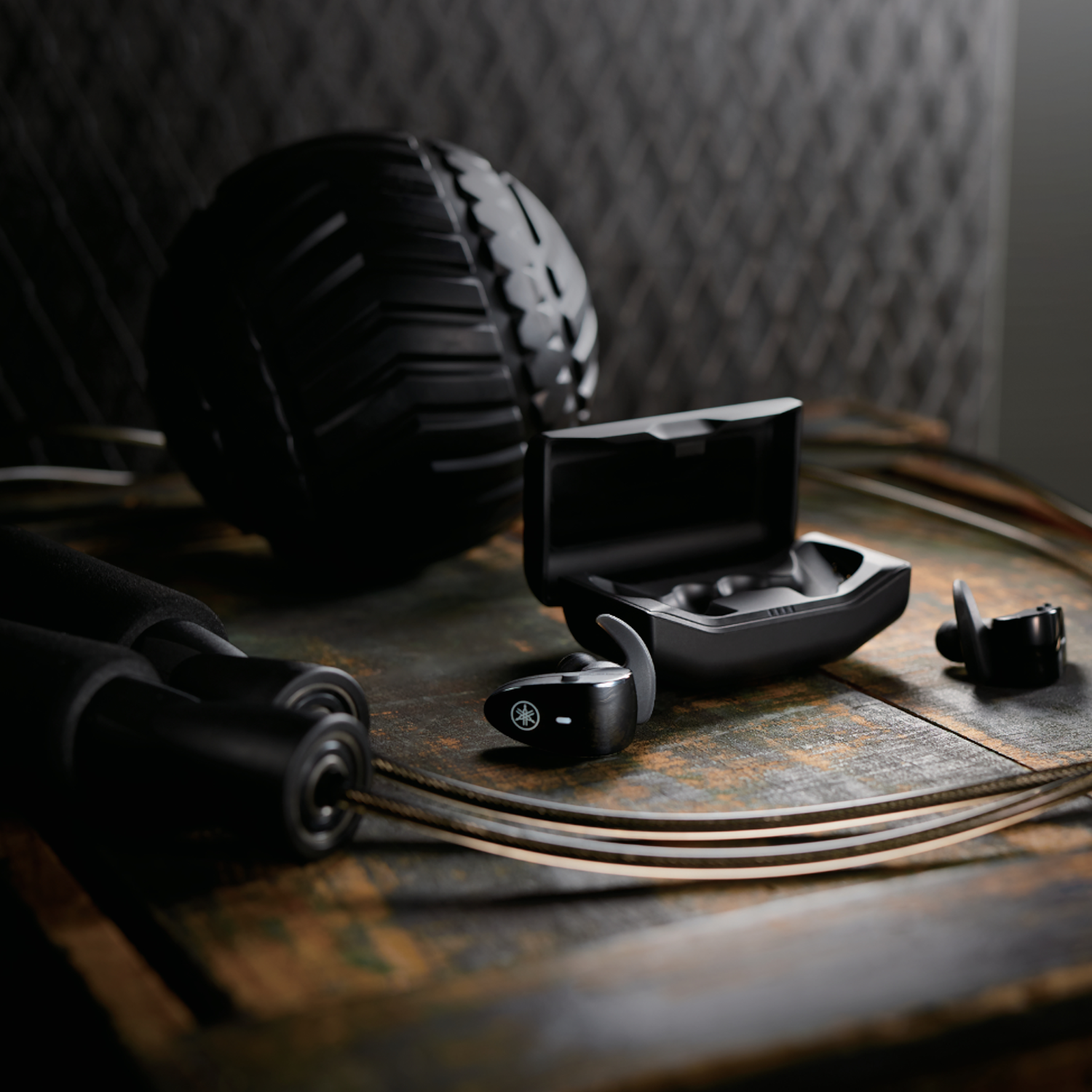 Auriculares negros en un estuche sobre una superficie de madera, con una pelota de masaje negra al fondo.