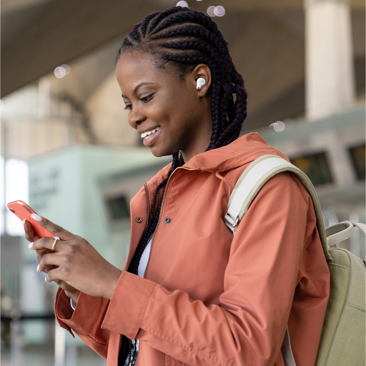 Kobieta z warkoczami, w słuchawkach, trzymająca telefon. Tło lotniska. Uśmiechnięta.