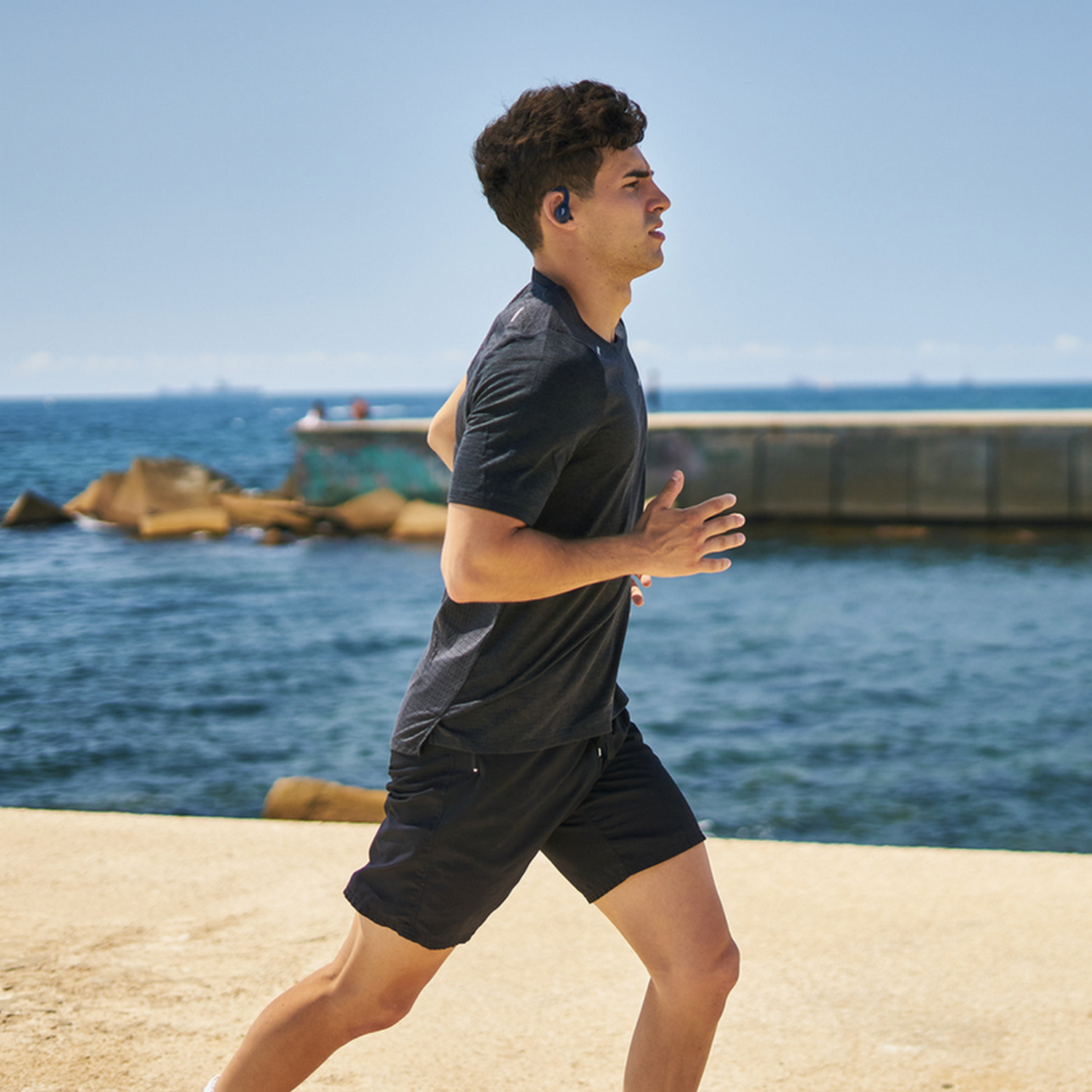 Un uomo che corre sulla spiaggia, indossa pantaloncini neri, una maglietta nera e auricolari.