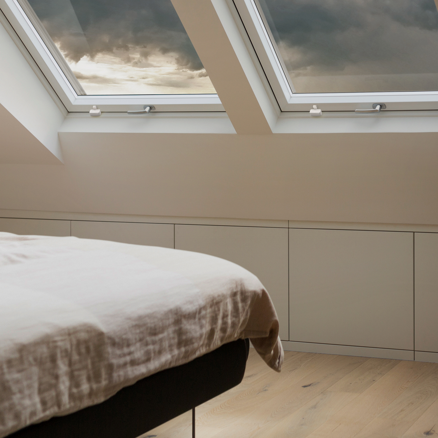 Sypialnia z dwoma oknami dachowymi, pokazującymi zachmurzone niebo. Poniżej neutralne kolorystycznie szafki i łóżko.