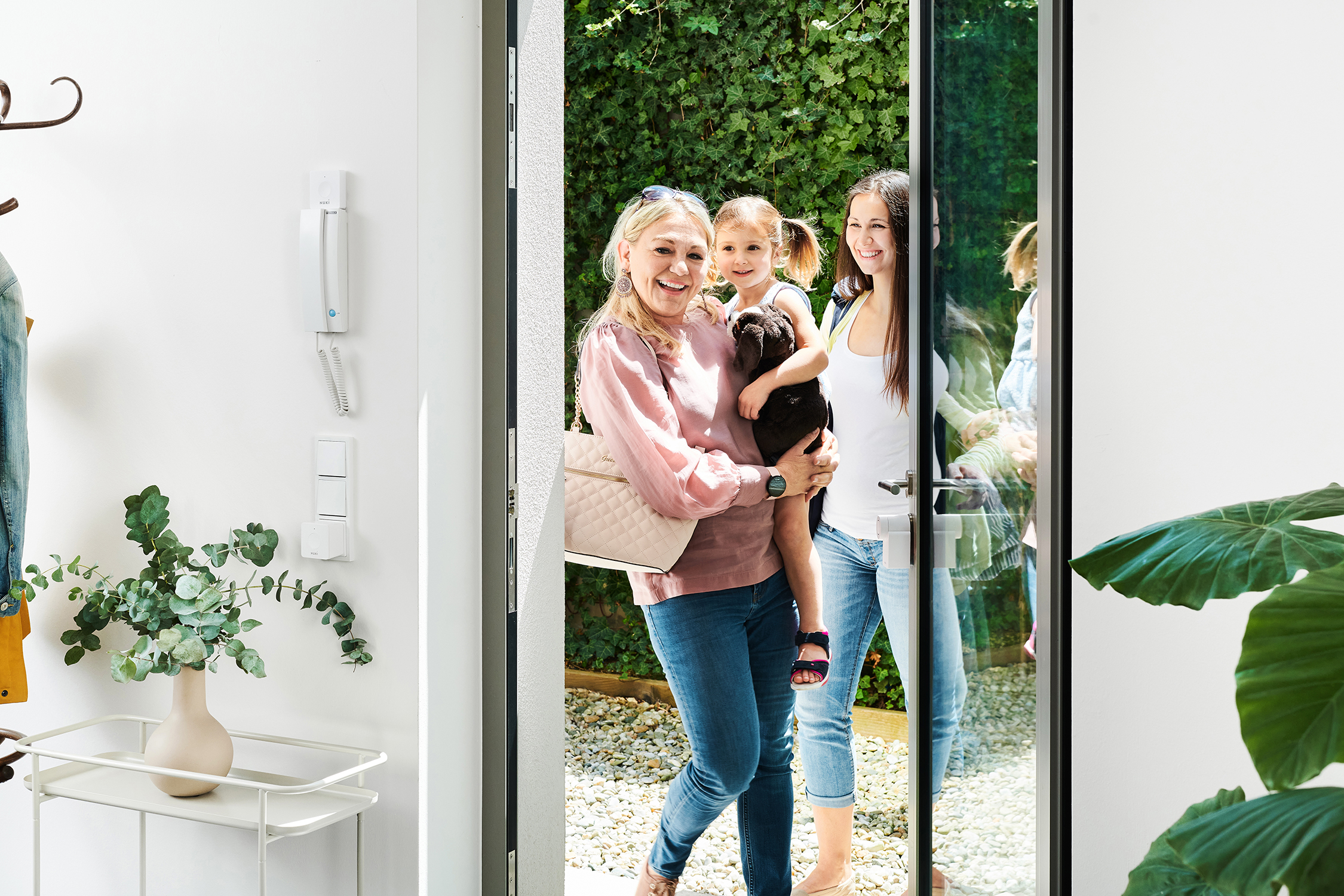 Rodzina w drzwiach. Matka trzyma dziecko, inna kobieta się uśmiecha. Widoczne zielone liście.