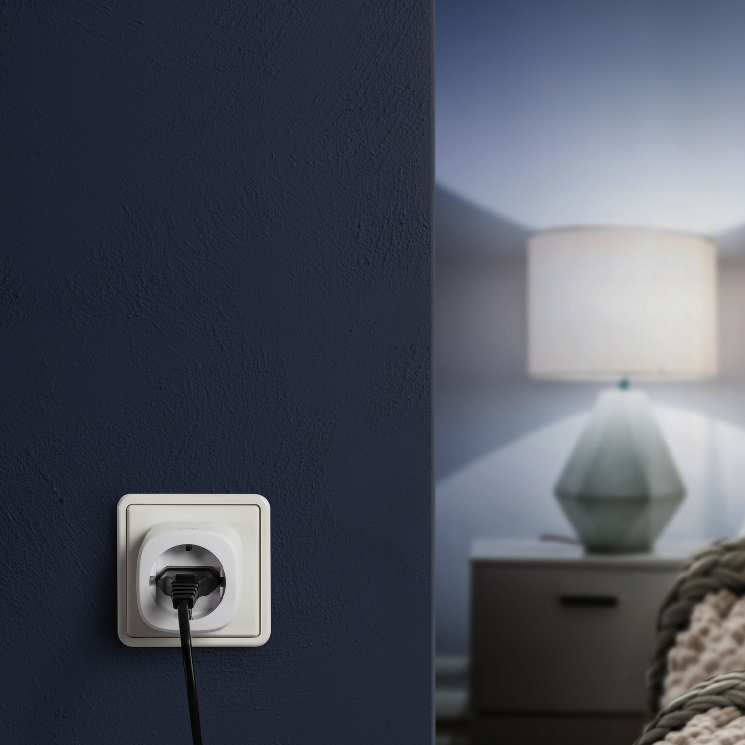 Weißer Smart Plug in einer dunkelblauen Steckdose. Eine Lampe und ein Nachttisch im Hintergrund.