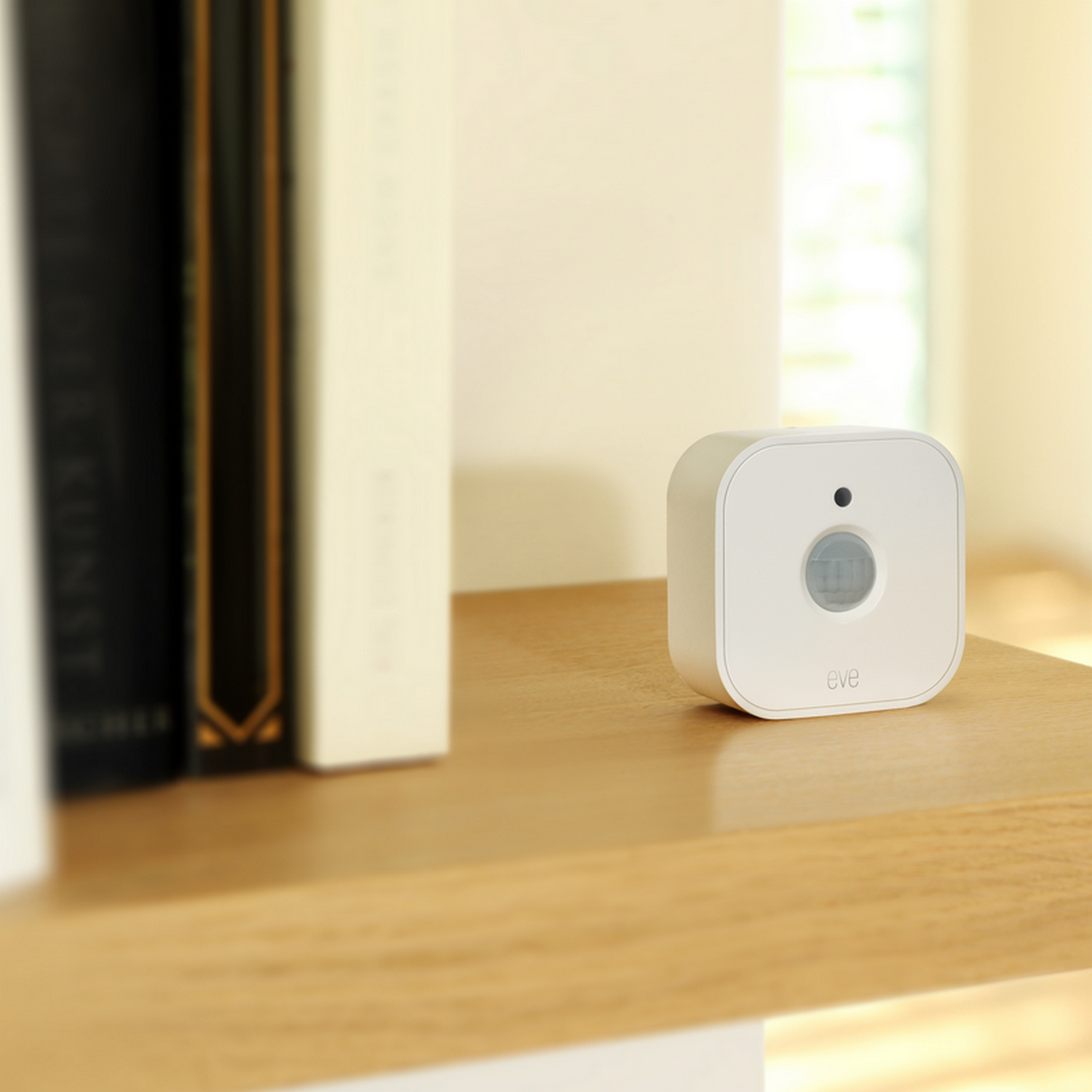Biały, kwadratowy czujnik smart home. Na drewnianej półce z książkami. Napis 'eve'.