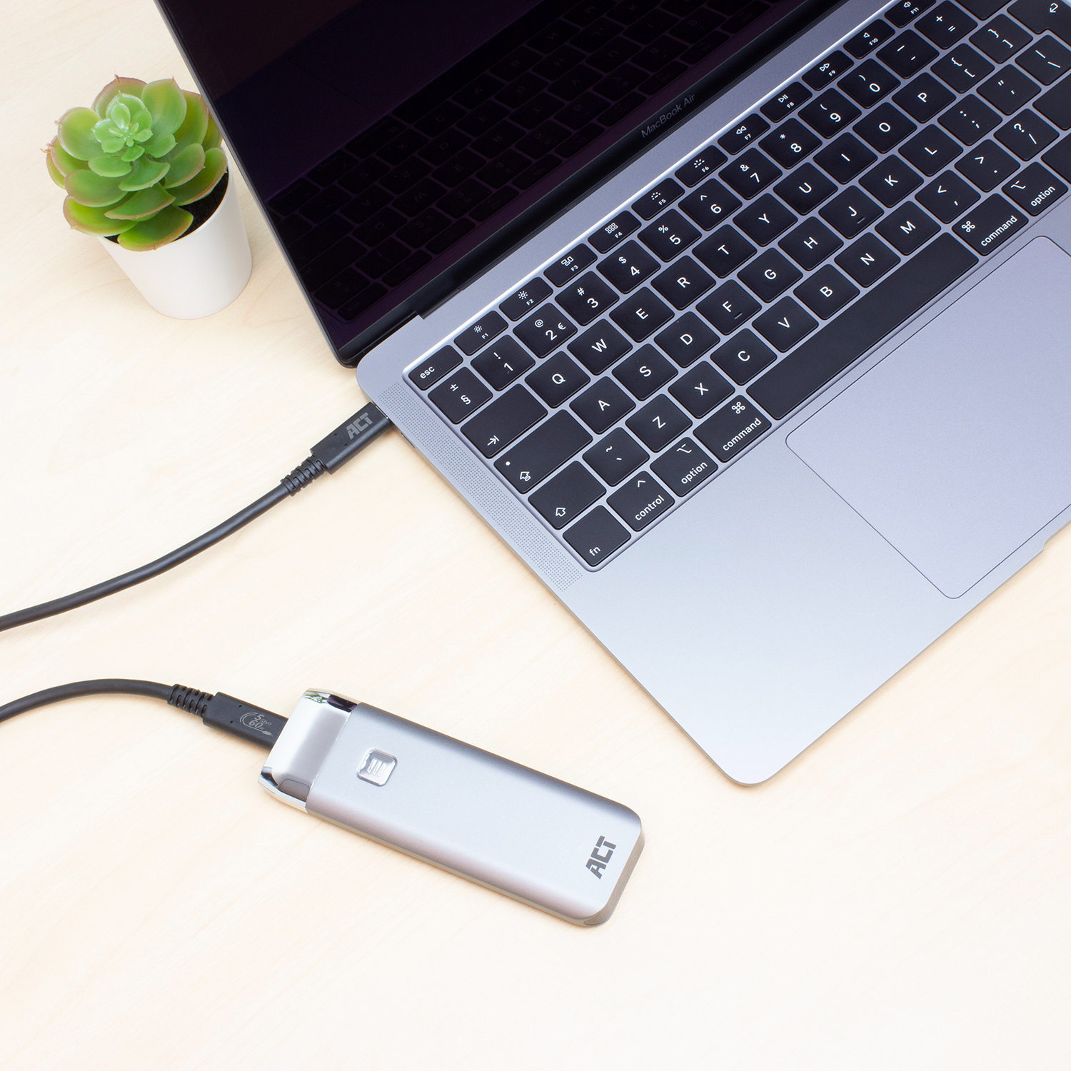 Een laptop verbonden met een zilveren apparaat via een zwarte kabel. Er staat een kleine plant op de tafel.