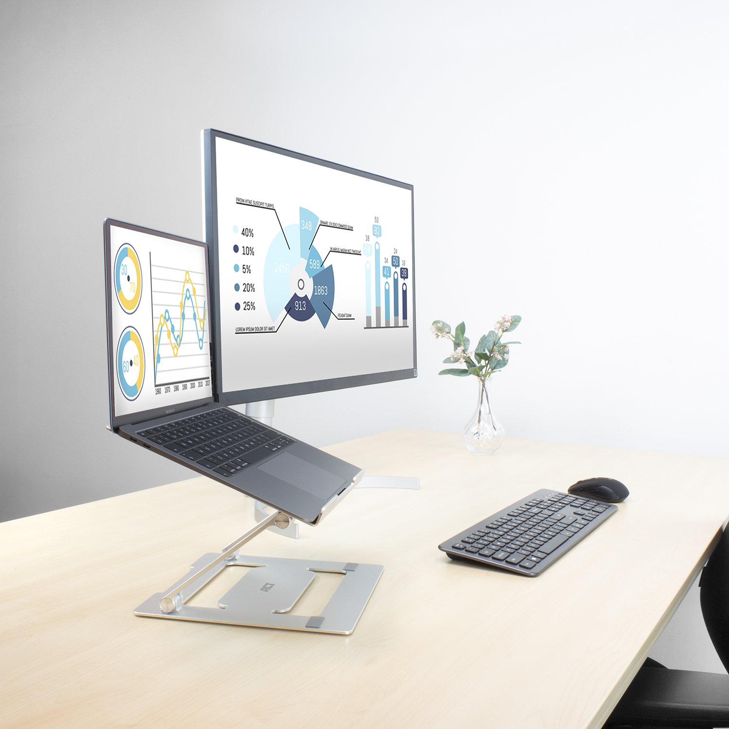 Laptop na stojaku podłączony do monitora. Klawiatura i mysz na stole. Wazon z kwiatami.
