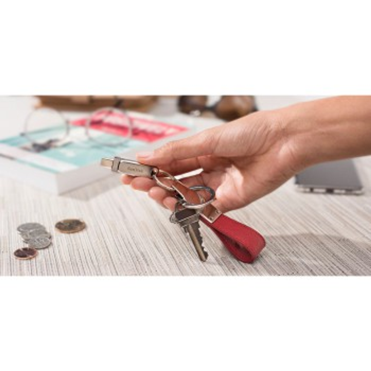 Hand holding keys and USB cable, coins, glasses, book on table.