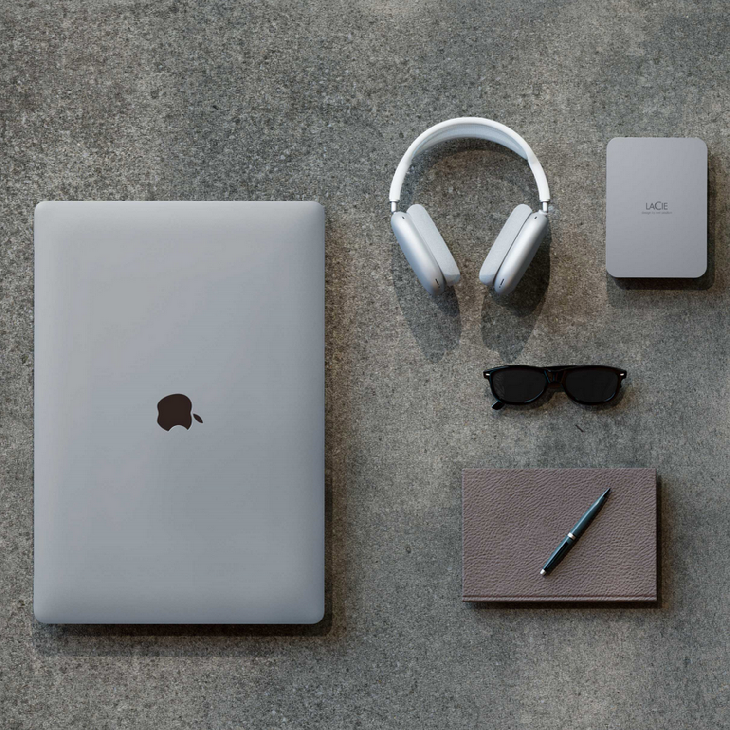 Widok z góry na zamkniętego szarego laptopa, słuchawki, dysk twardy, okulary, długopis i notatnik na szarej powierzchni.