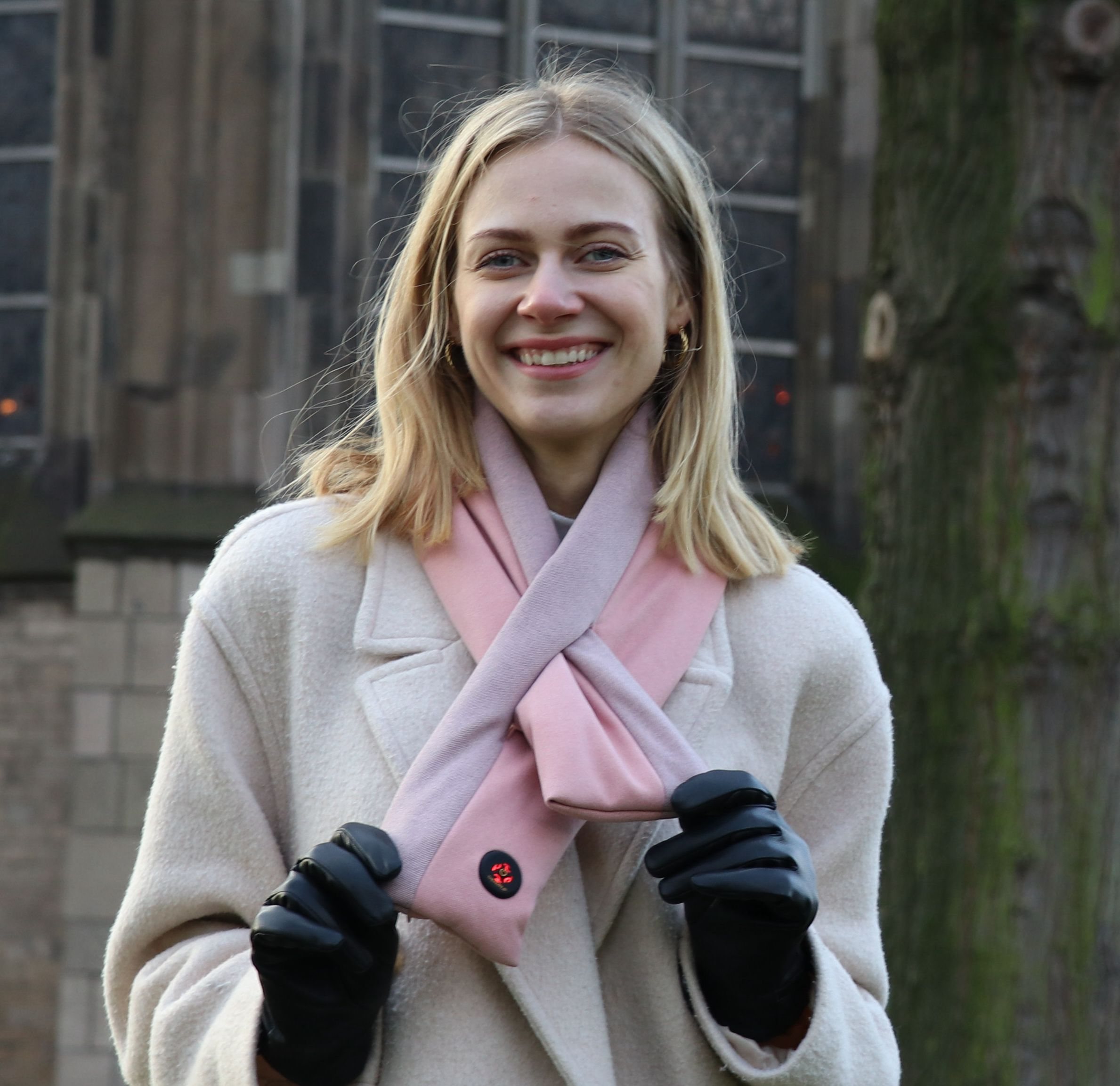 Frau mit blonden Haaren lächelt, trägt einen weißen Mantel, einen rosa Schal und schwarze Handschuhe.