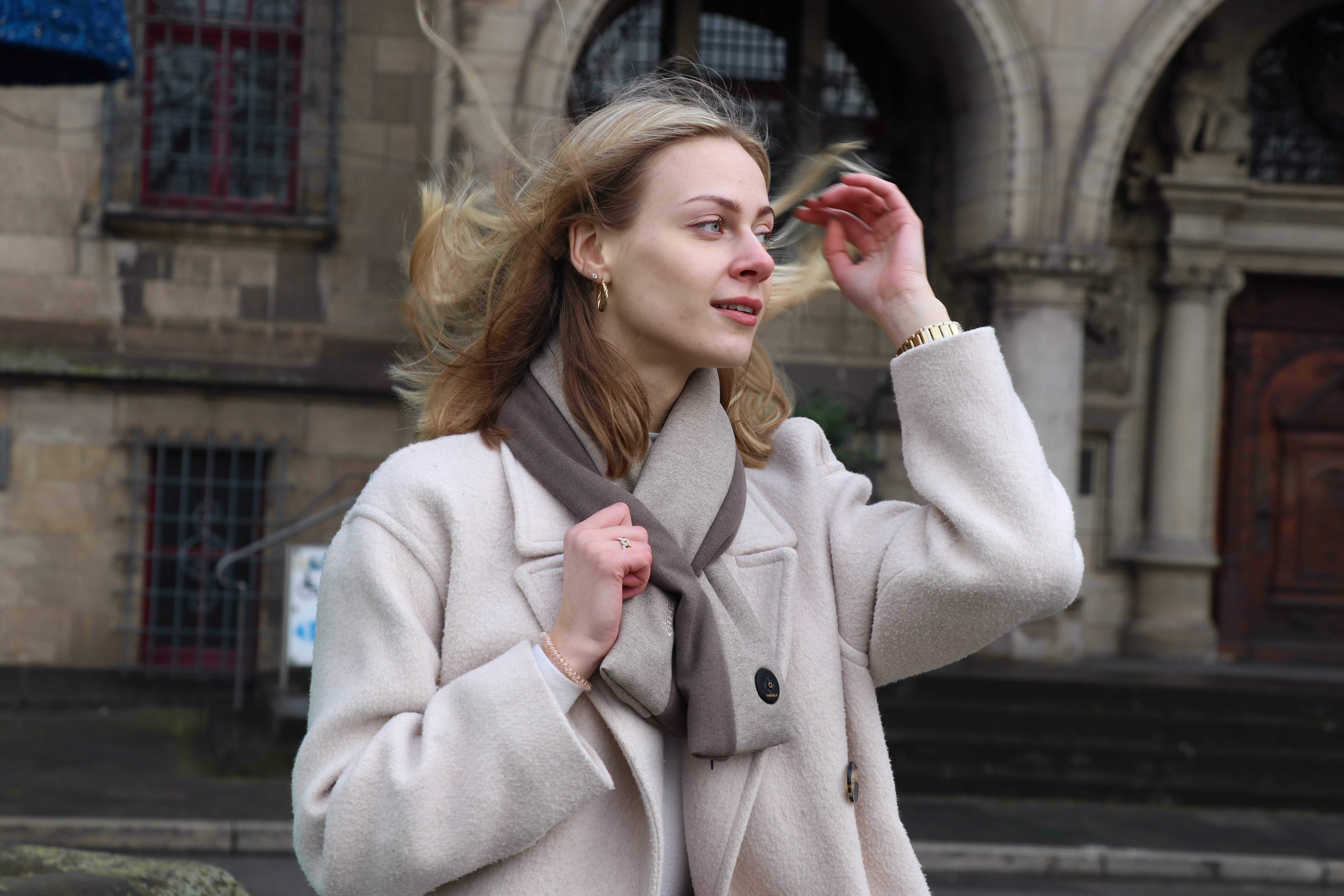 Eine blonde Frau mit weißem Mantel und Schal steht vor einem Gebäude. Wind weht durch ihre Haare.