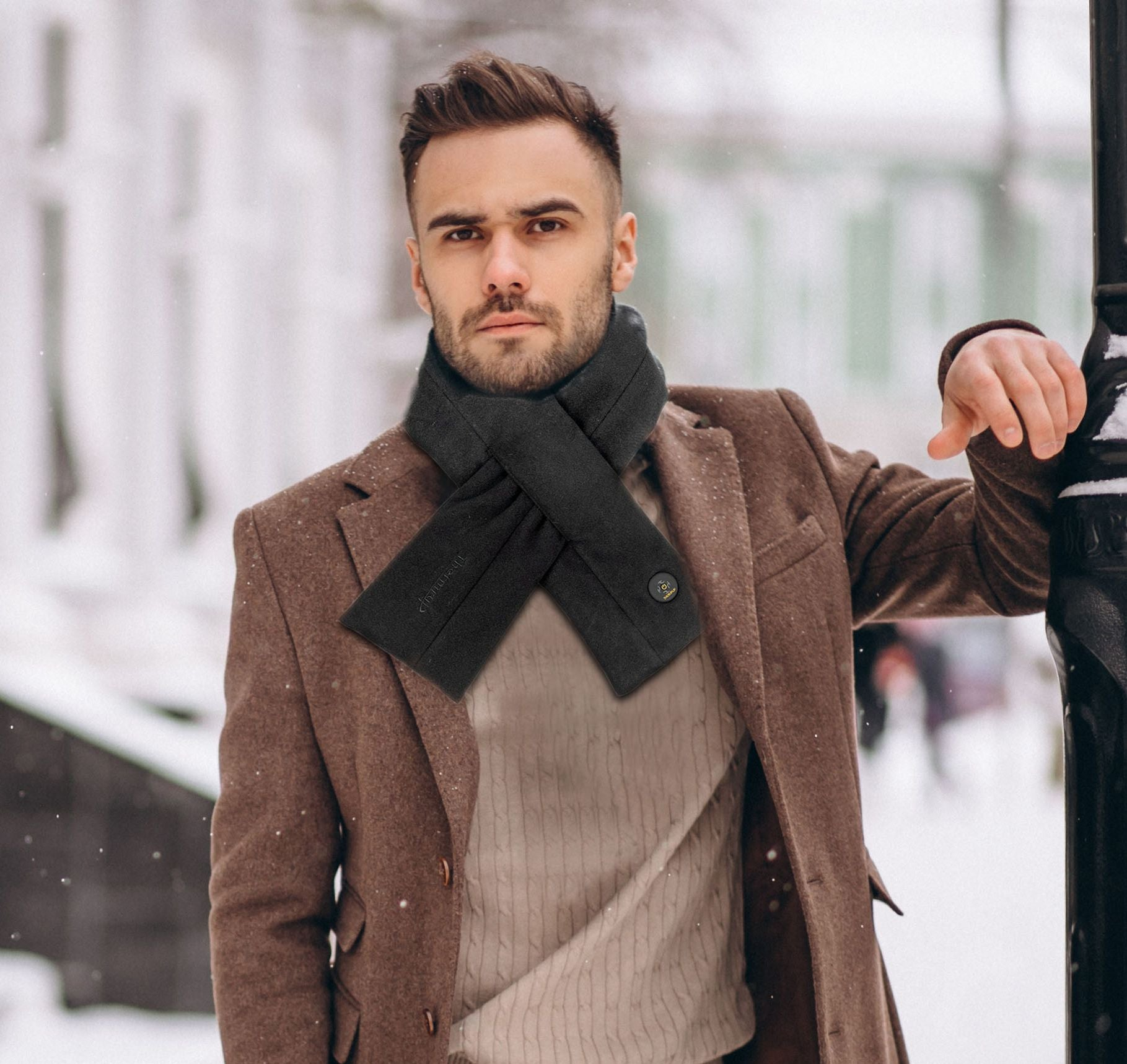 Mann in braunem Mantel und schwarzem Schal, steht im Schnee und lehnt an einer Metallstange. Winterszene.