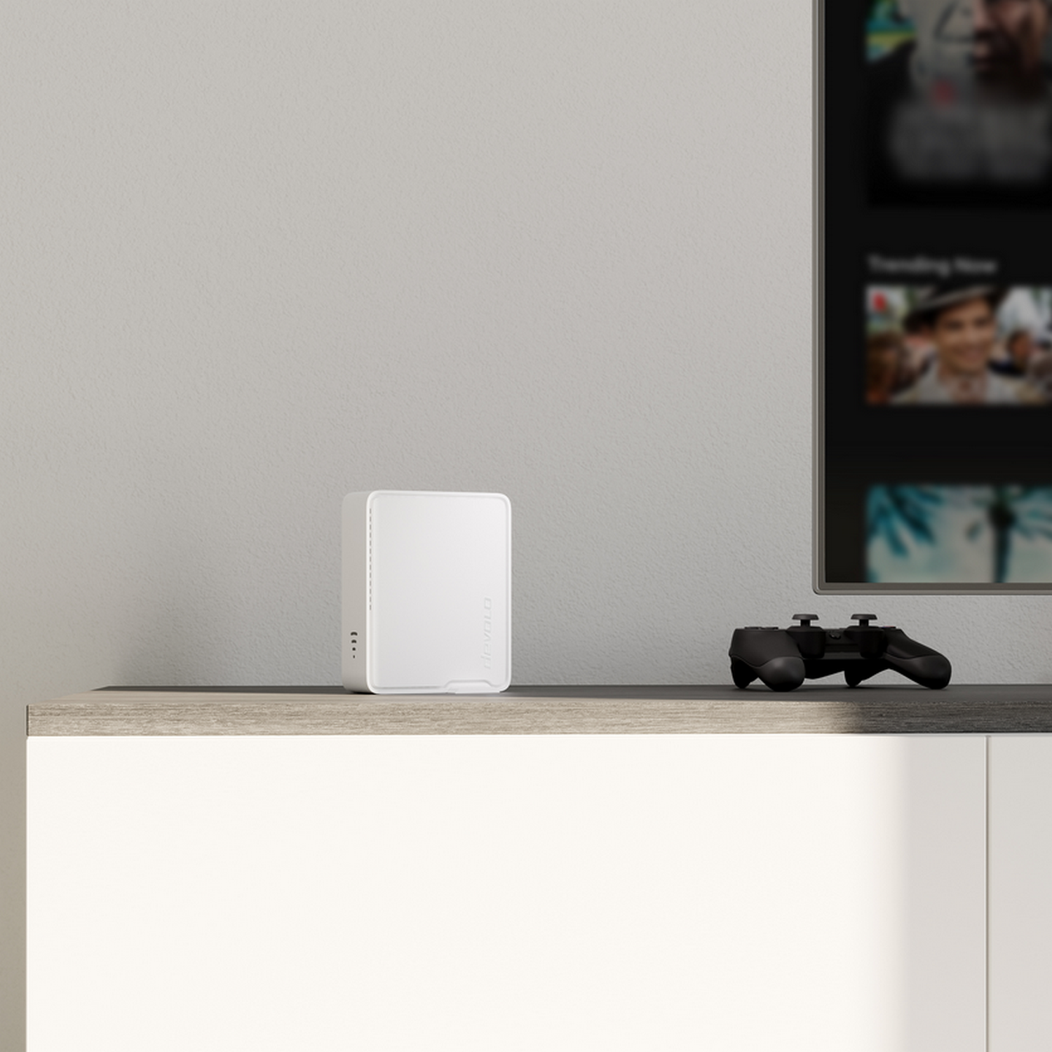 Białe urządzenie multimedialne na półce obok kontrolera do gier. Telewizor z rozmytym interfejsem w tle.