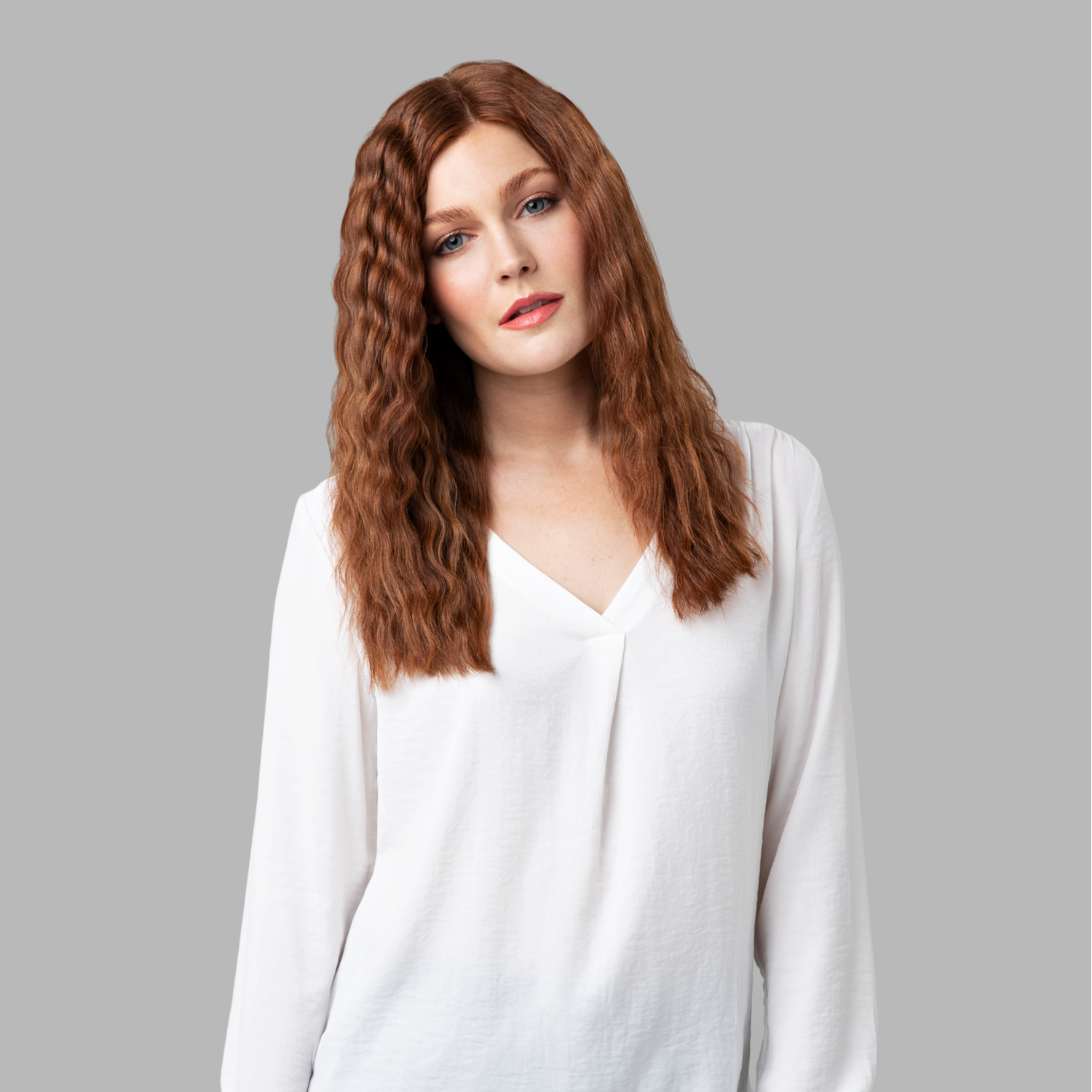 Ruda kobieta w białej bluzce z dekoltem w serek. Szare tło. Patrzy w kamerę.
