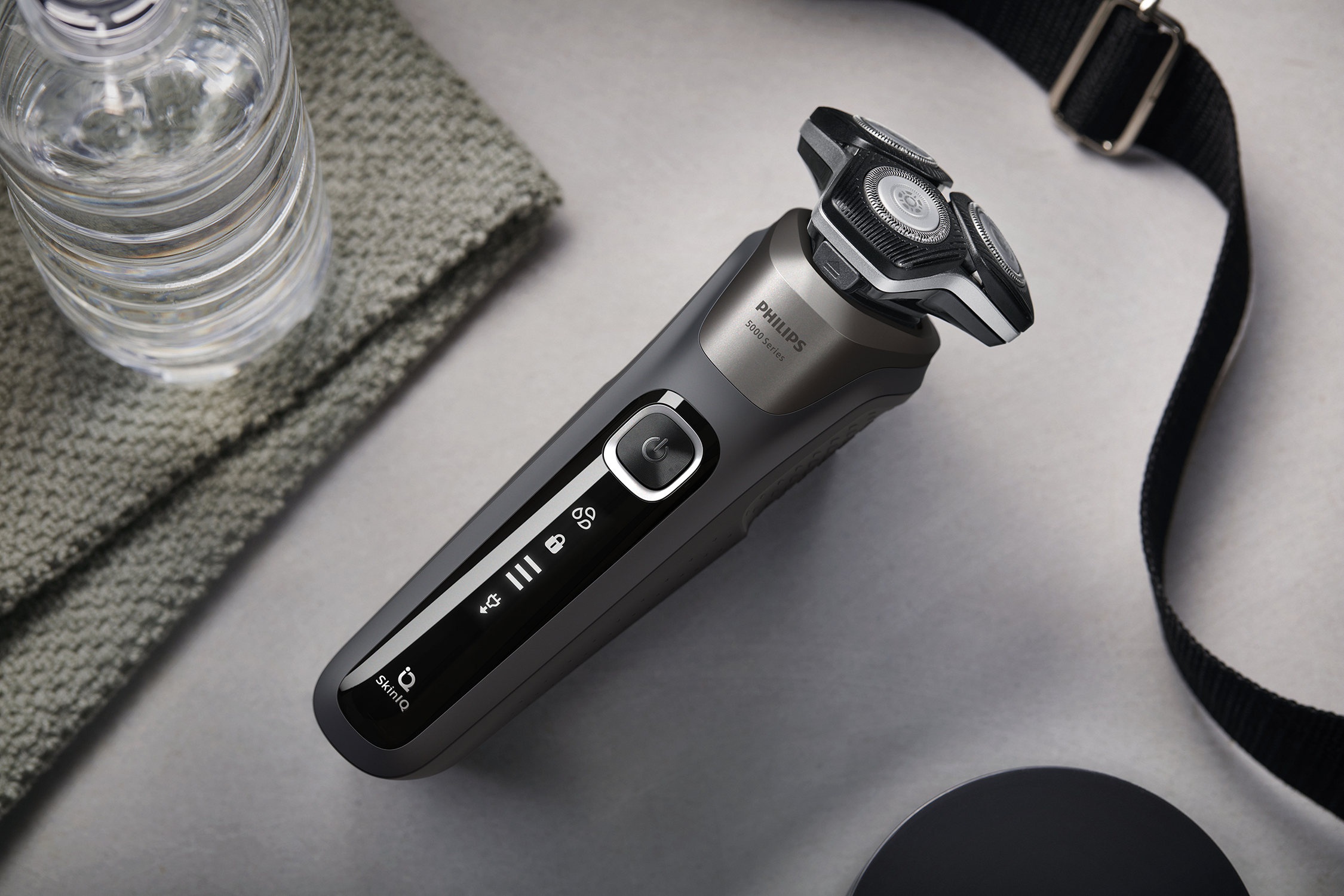 A Philips shaver on a surface with a water bottle, towel, and strap. The shaver is black and gray.
