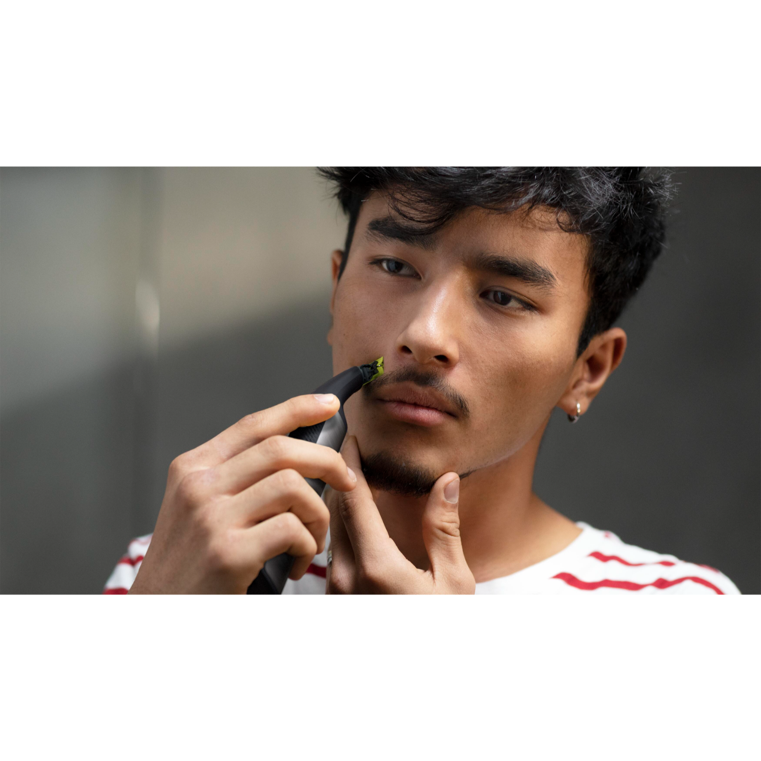 A man is trimming his mustache with a black trimmer. He wears a white shirt.