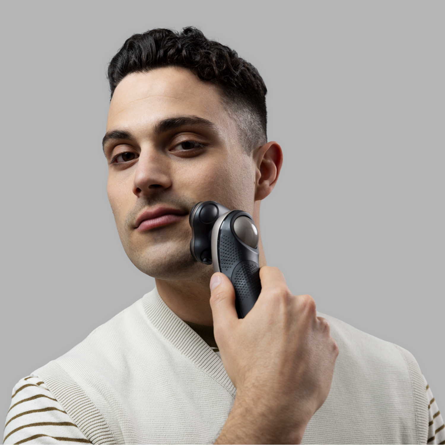 Uomo che si rade con rasoio elettrico. Sfondo grigio, maglione chiaro. Focalizzato sul viso.