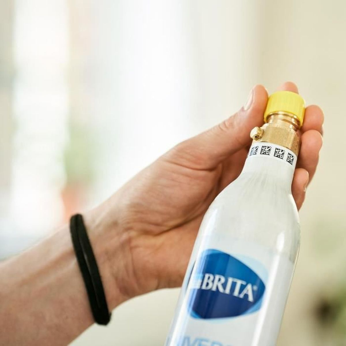 Eine Hand hält eine weiße Brita-Wasserfilterflasche mit gelbem Verschluss und schwarzem Armband am Handgelenk.