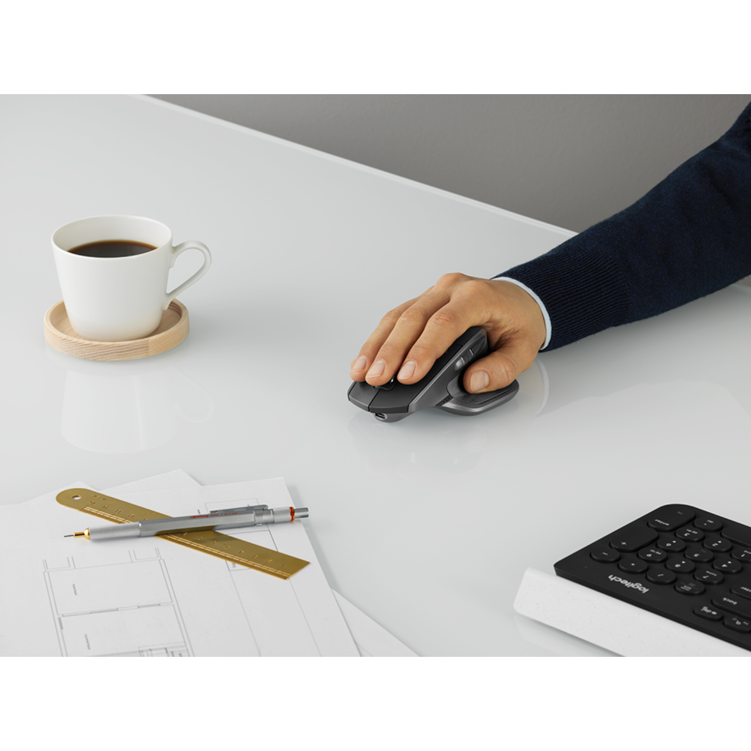 Osoba używająca myszy pionowej na białym biurku. Na biurku również: filiżanka kawy, linijka i klawiatura.