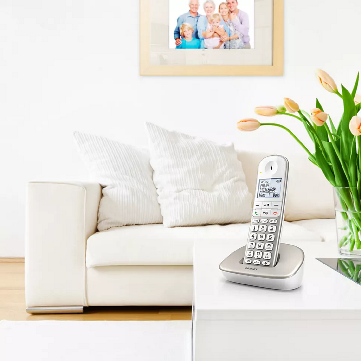 Un téléphone sans fil blanc est sur une table avec un vase de fleurs. Une photo de famille est accrochée au mur.