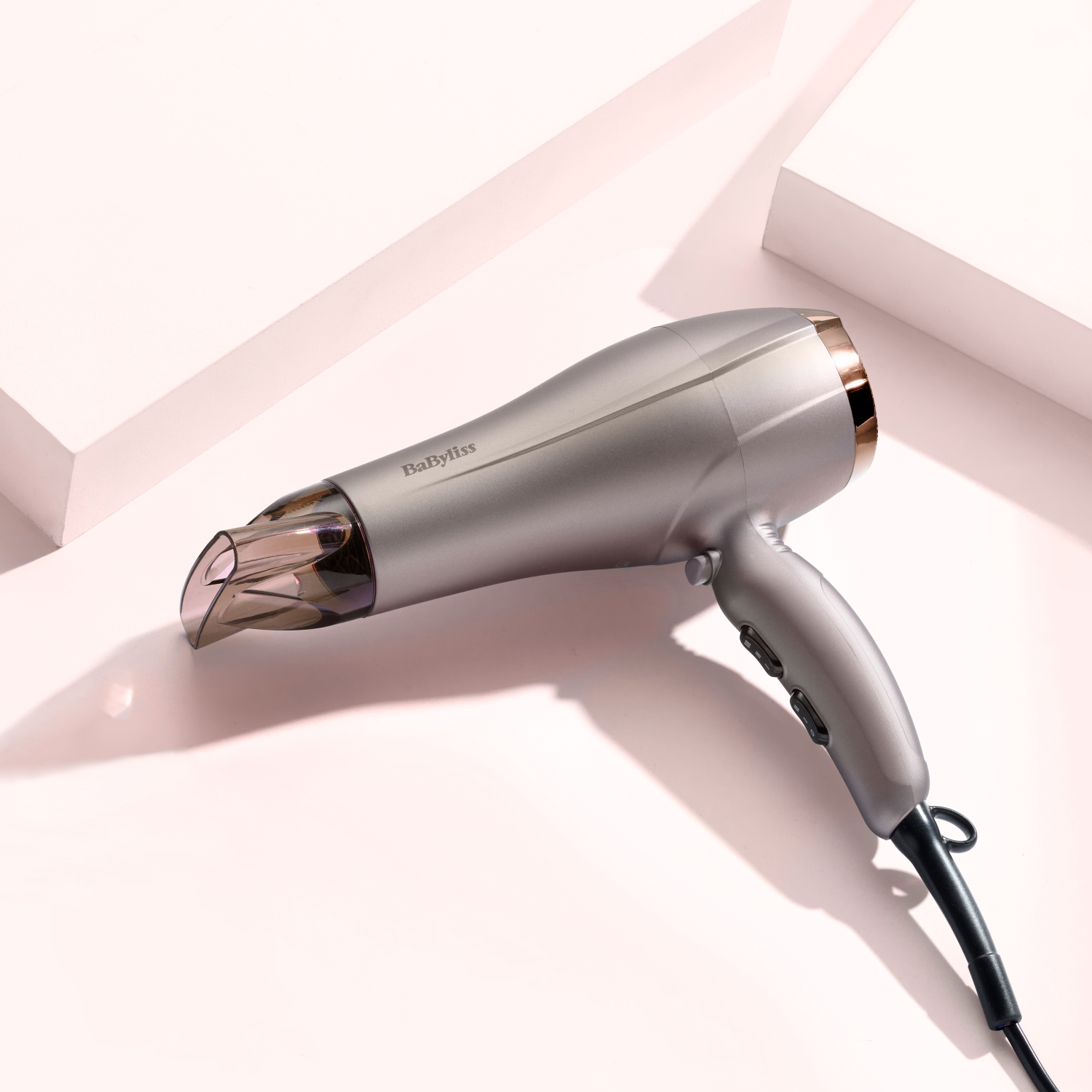 Un asciugacapelli BaByliss argento e oro su sfondo rosa con forme geometriche.