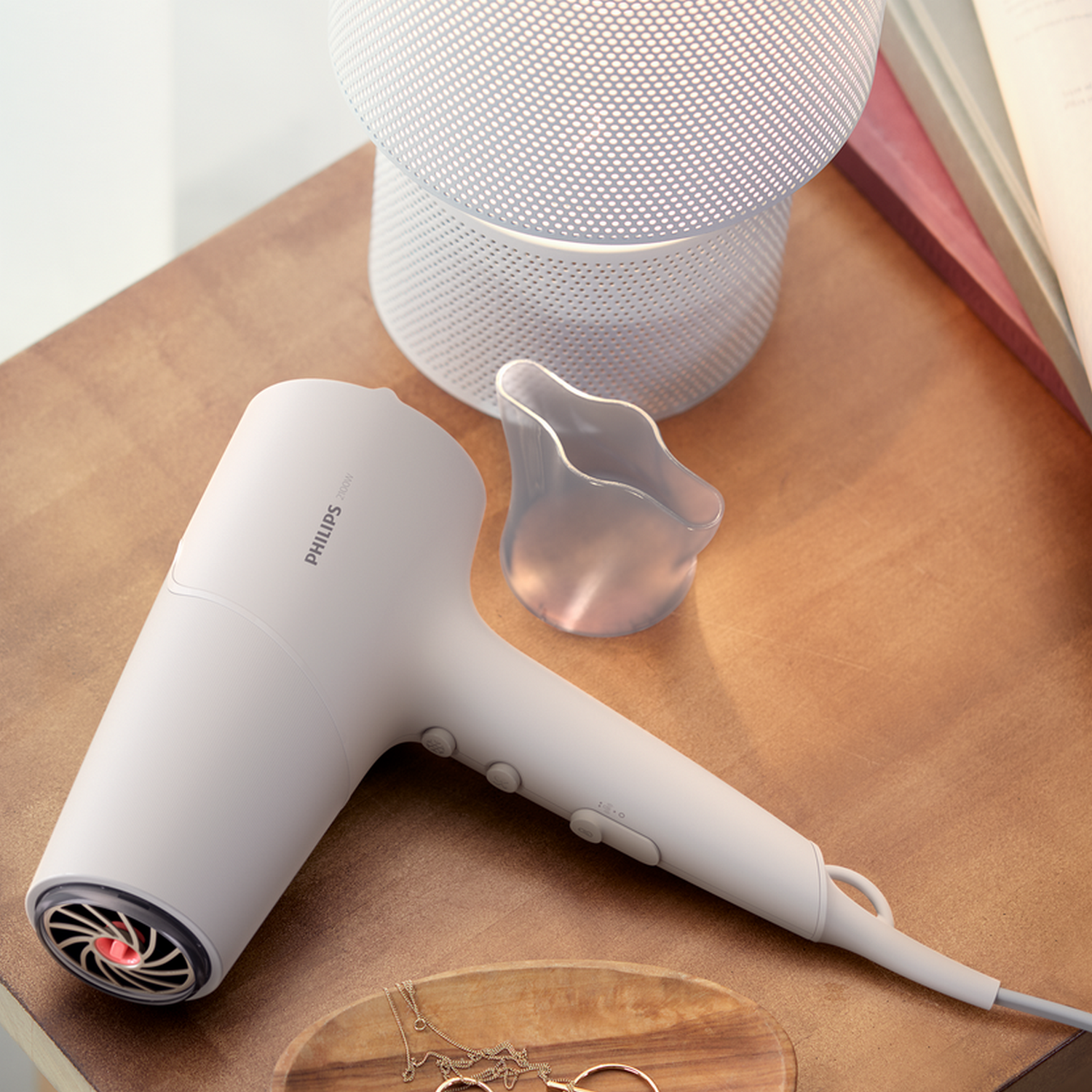 Biała suszarka do włosów na drewnianym stole z lampą, naszyjnikiem i przezroczystym plastikiem.