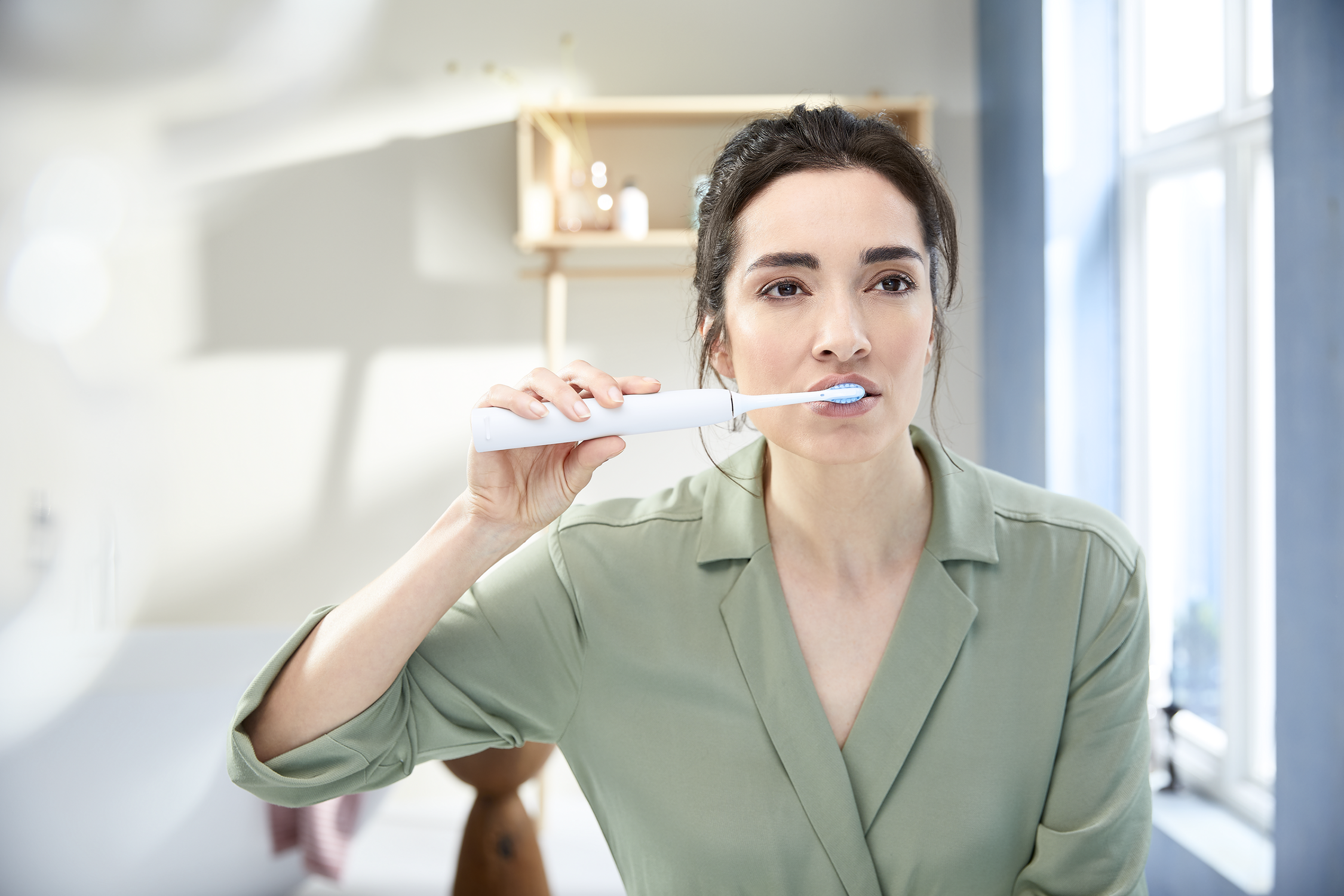 Mujer cepillándose los dientes con un cepillo eléctrico blanco en un baño.
