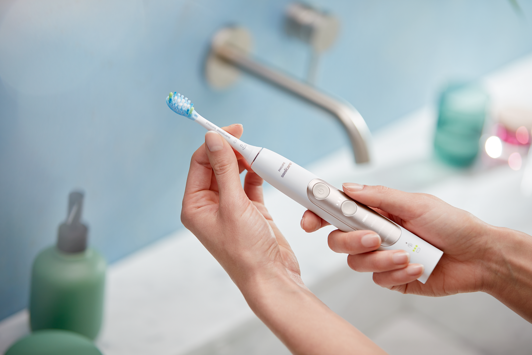 Persona sosteniendo un cepillo de dientes eléctrico blanco junto a un lavabo, fondo azul y blanco.