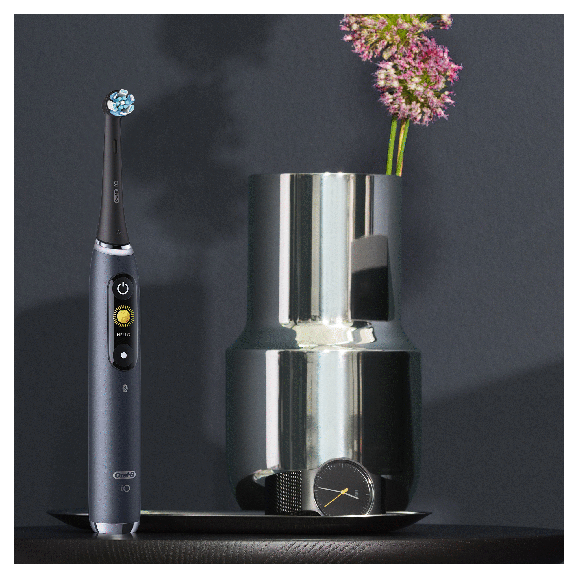 Szczoteczka elektryczna, wazon z kwiatami, zegarek na tacy. Wszystko na ciemnym tle.