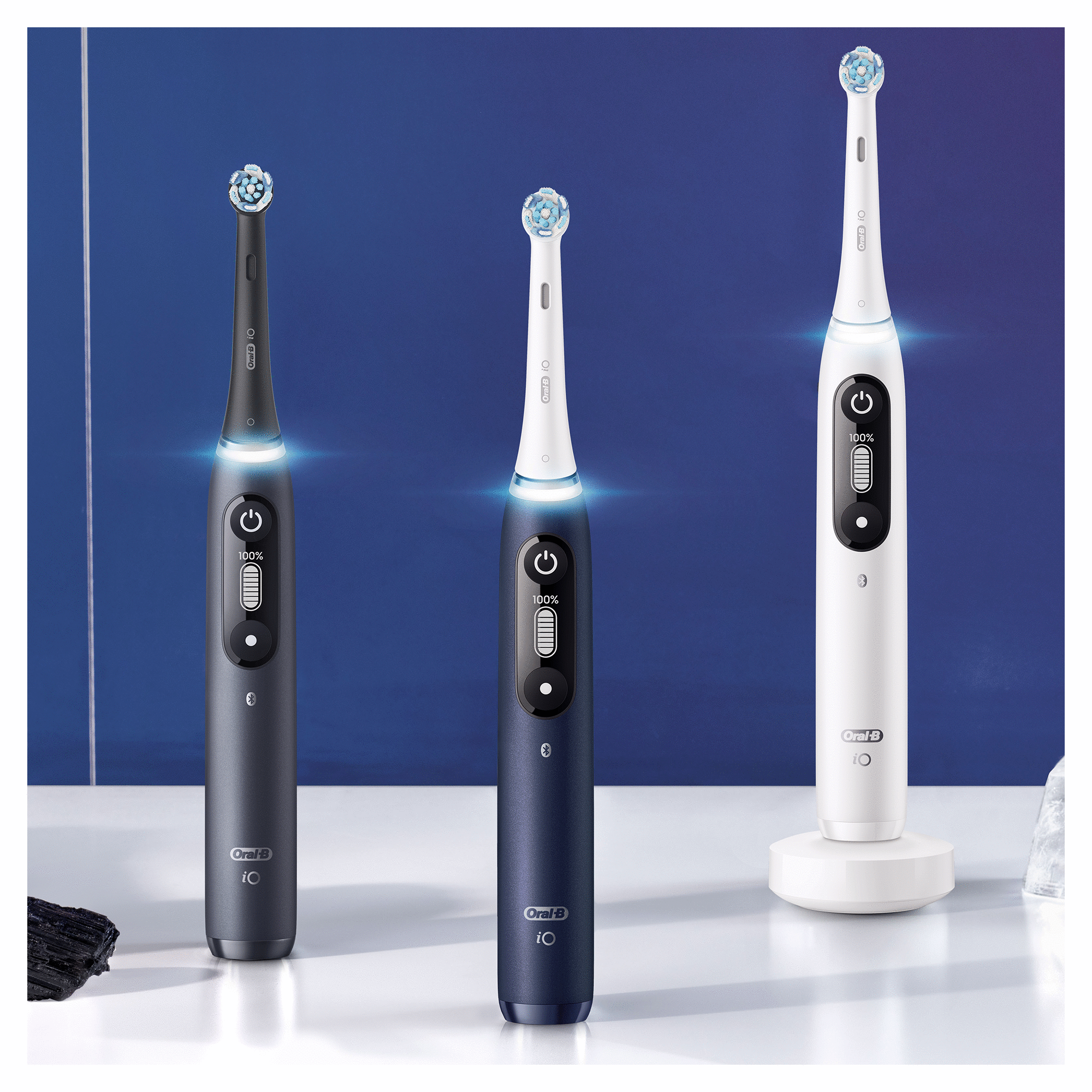 Tre spazzolini elettrici, nero, blu e bianco, con testine illuminate e una base di ricarica su sfondo blu.