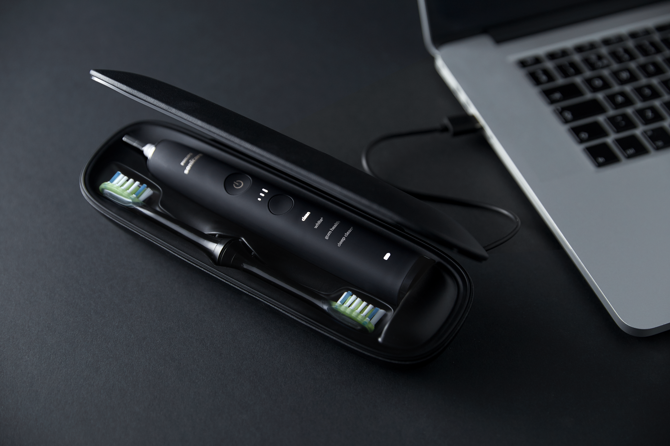 Czarna elektryczna szczoteczka do zębów w etui obok laptopa. Szczoteczka się ładuje.