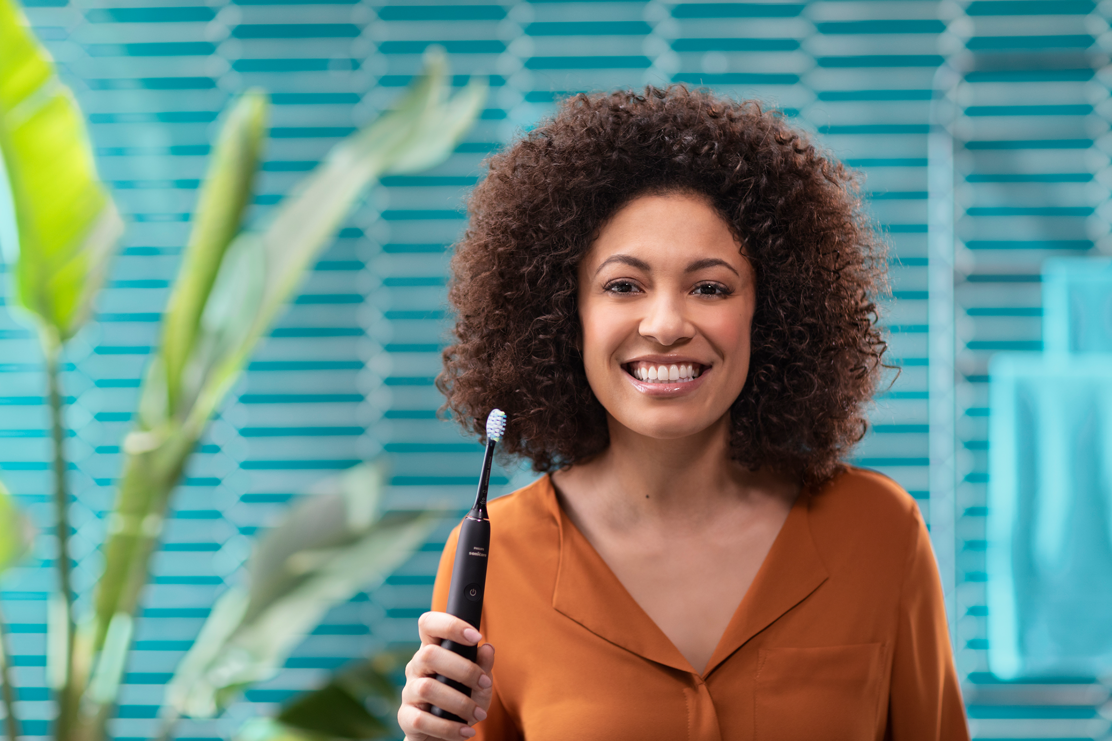 Kobieta z kręconymi włosami trzyma szczoteczkę do zębów i się uśmiecha. Tło jest niebieskie i zielone.