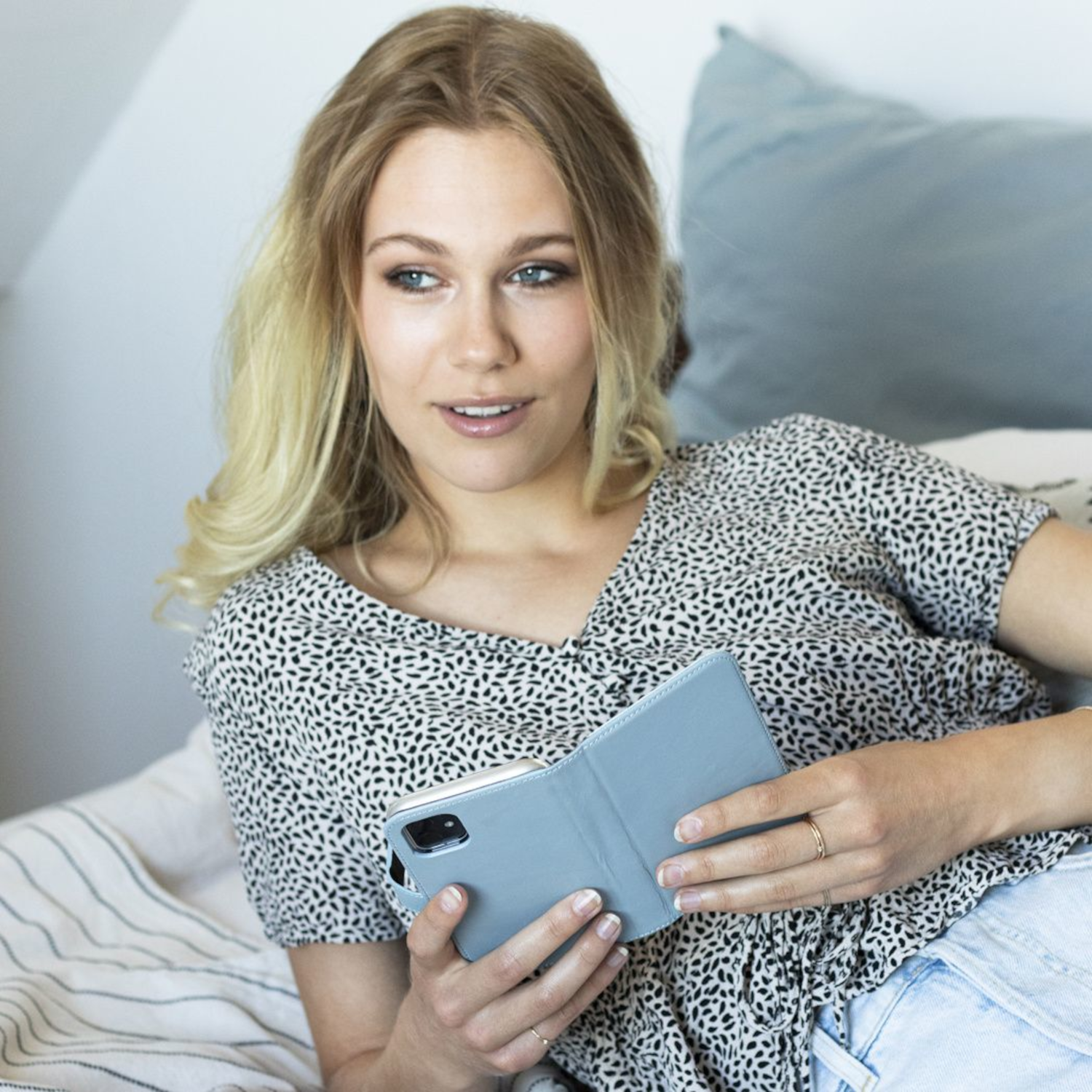 Blonde vrouw met een lichtblauwe telefoonhoes. Ze ligt op een bed met kussens.