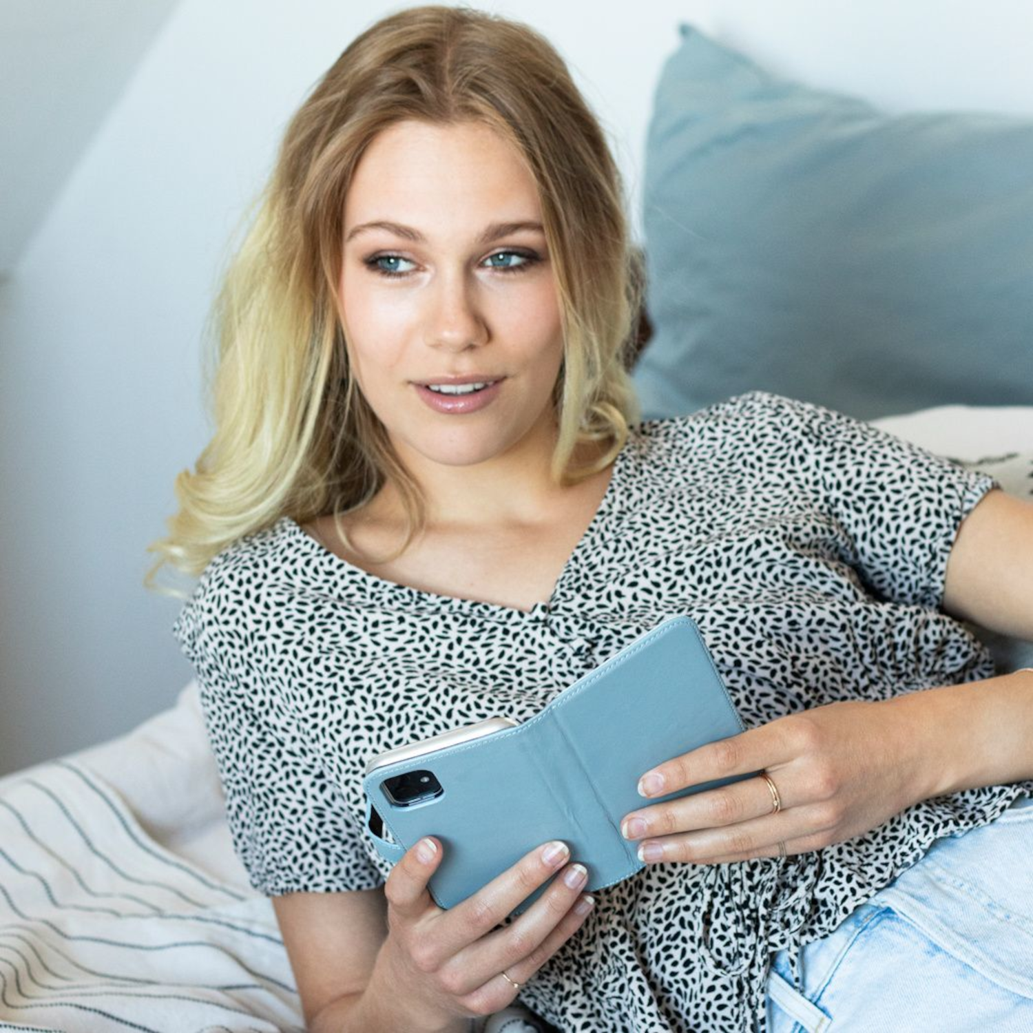 Vrouw houdt een blauwe telefoonhoes vast. Ze is blond, draagt een bloes, op een bed.