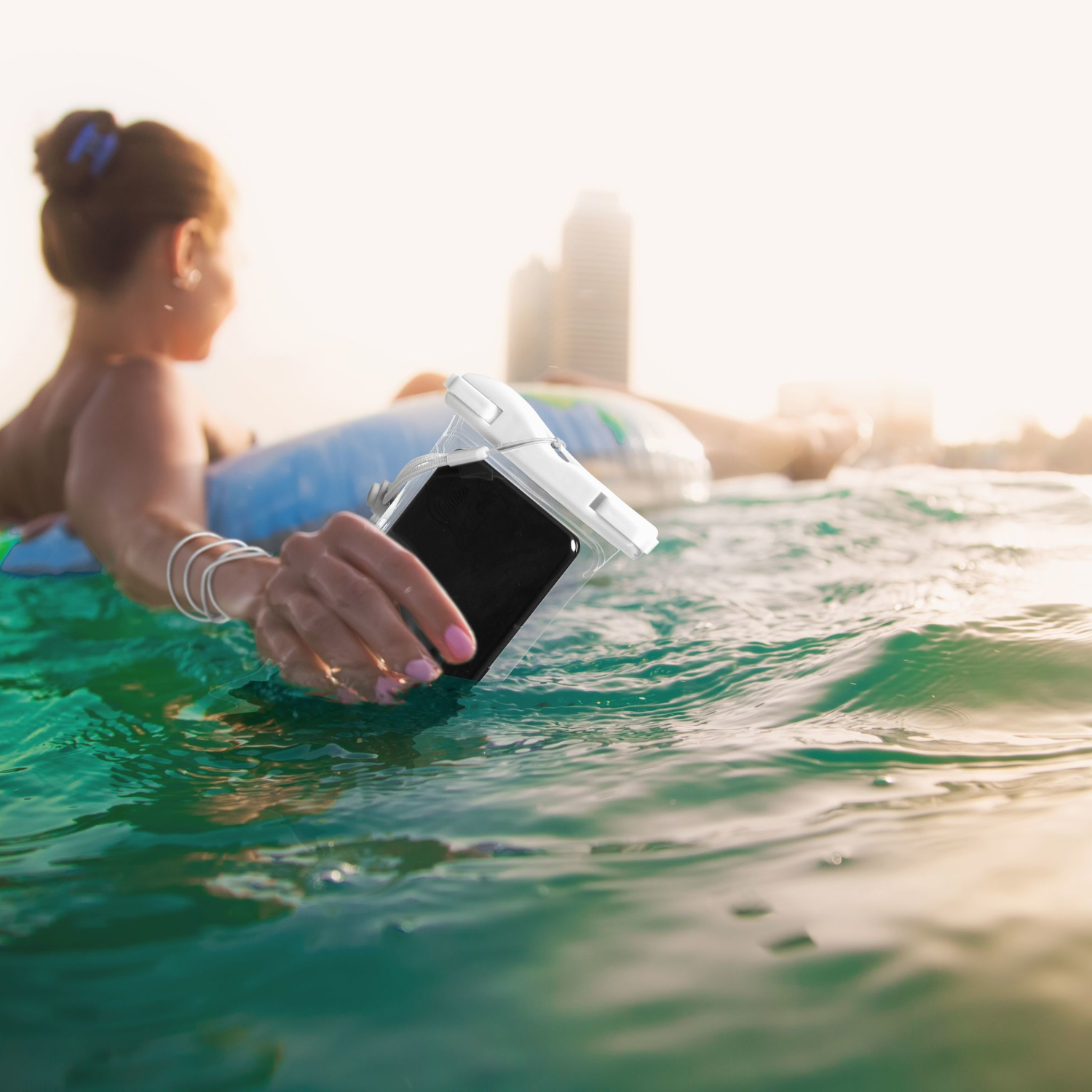 Donna in acqua che tiene il telefono in una custodia impermeabile. Lo sfondo mostra una spiaggia e degli edifici.