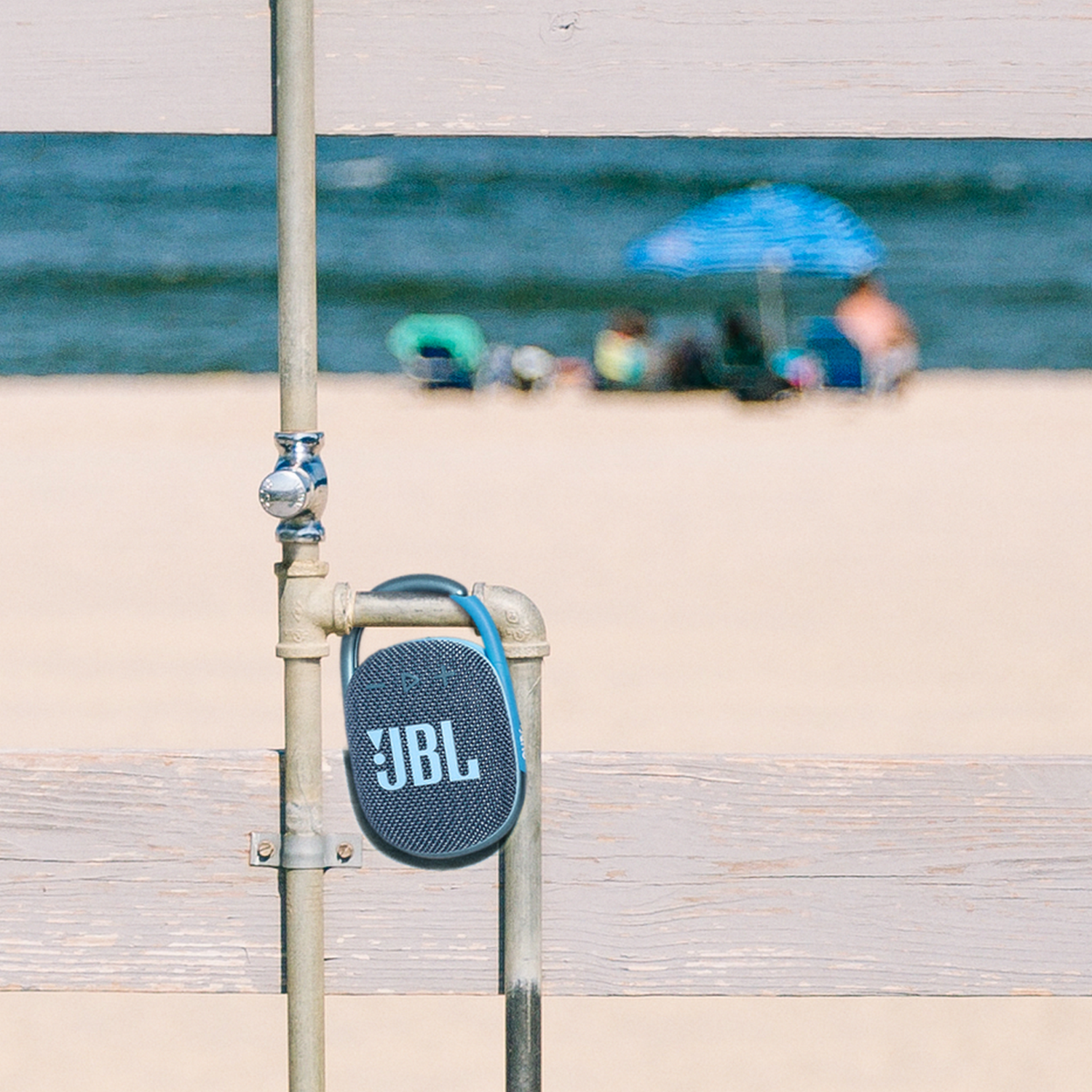 Mały niebieski głośnik wisi na metalowej rurze przy plaży. Scena na plaży jest w tle.