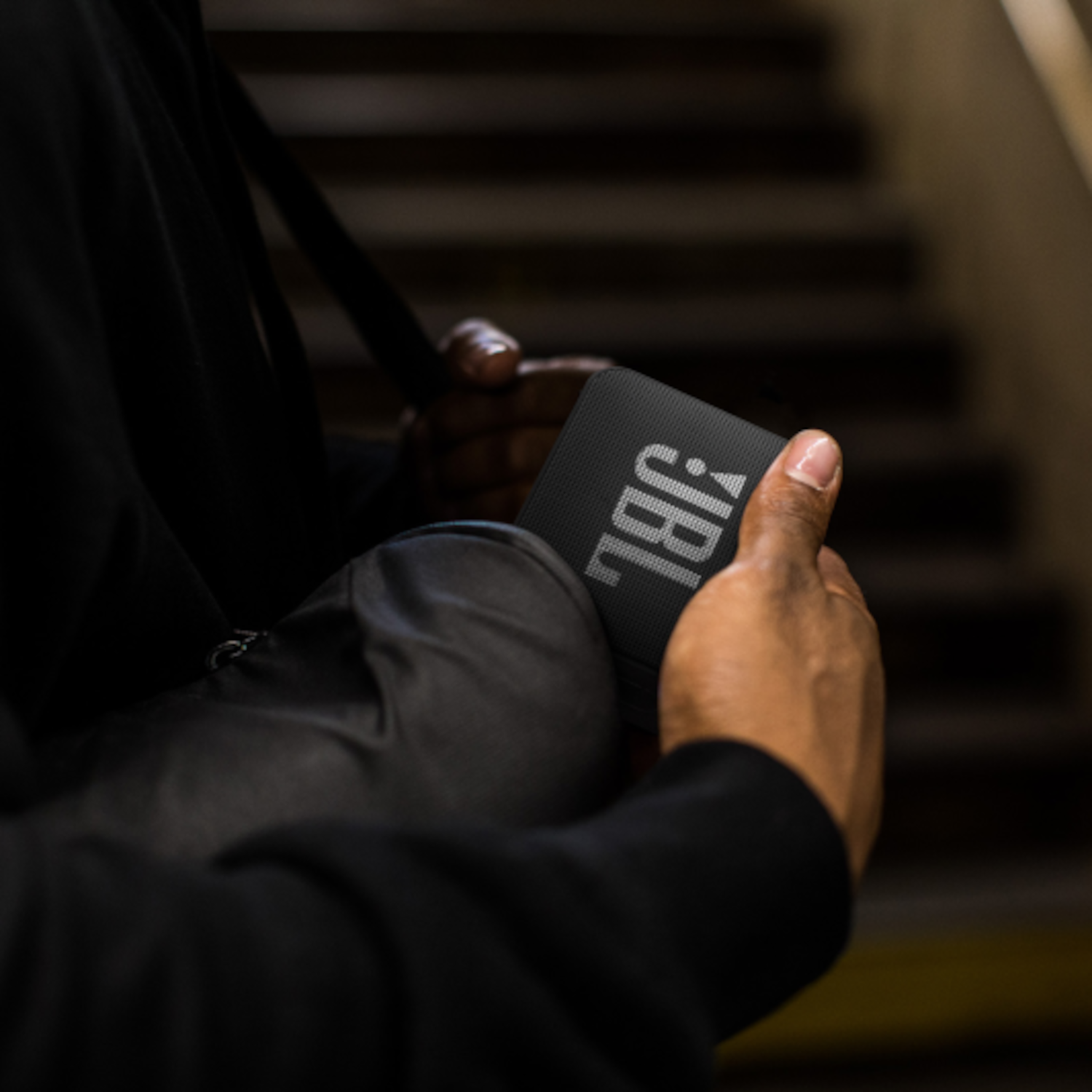 Person hält einen schwarzen JBL-Lautsprecher. Schwarze Jacke, Hintergrund mit Treppen.