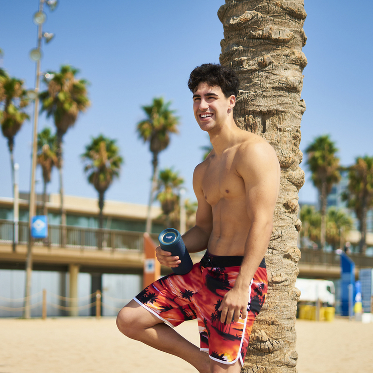 Un hombre está de pie junto a una palmera en la playa, sin camisa. Sostiene un altavoz azul y sonríe.