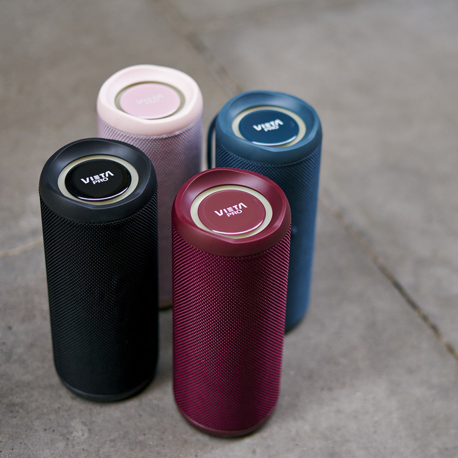 Cuatro altavoces cilíndricos en negro, rosa, granate y azul. Colocados sobre superficie de piedra.