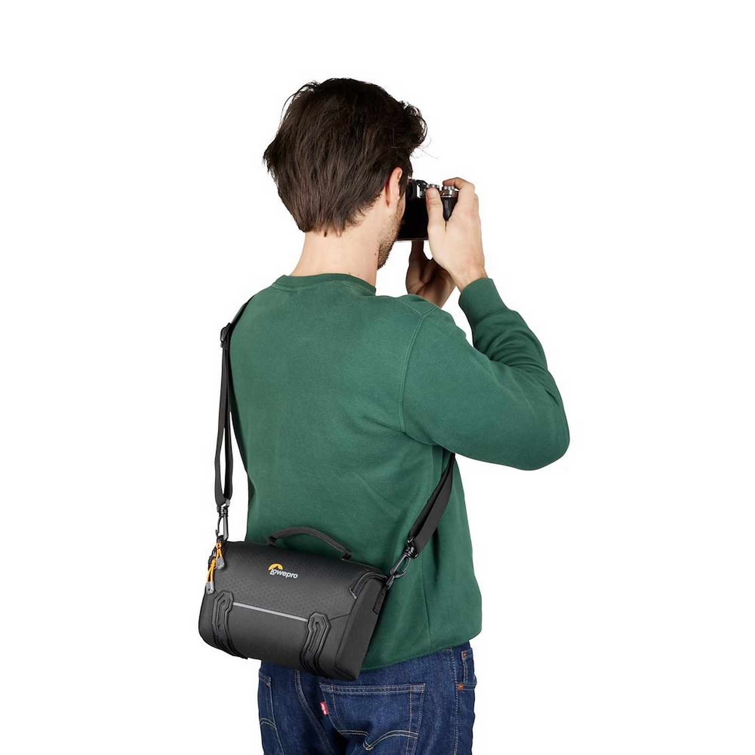 A man in a green sweater takes a photo with a camera and a black bag.