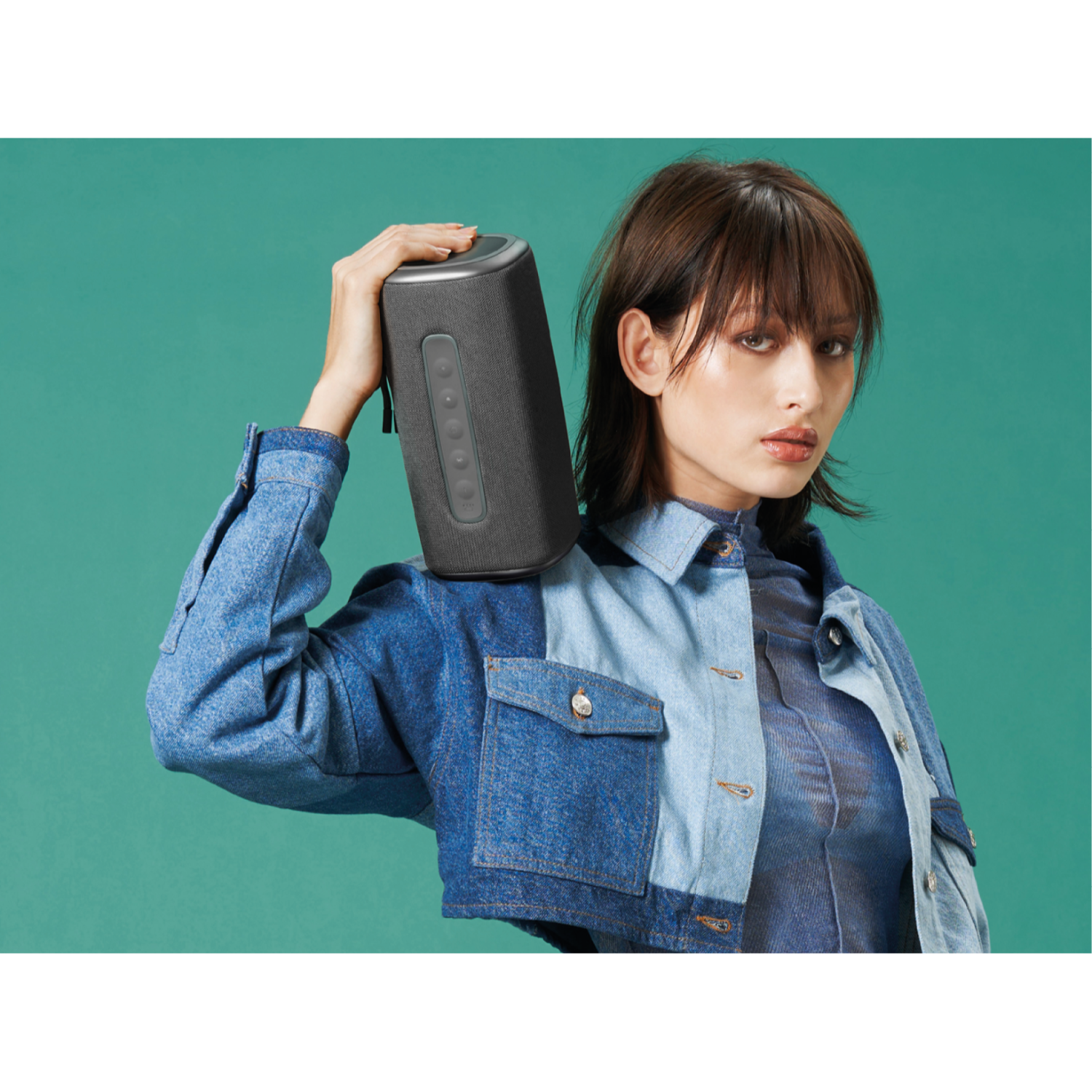 Kobieta trzymająca ciemny głośnik na ramieniu, turkusowe tło.