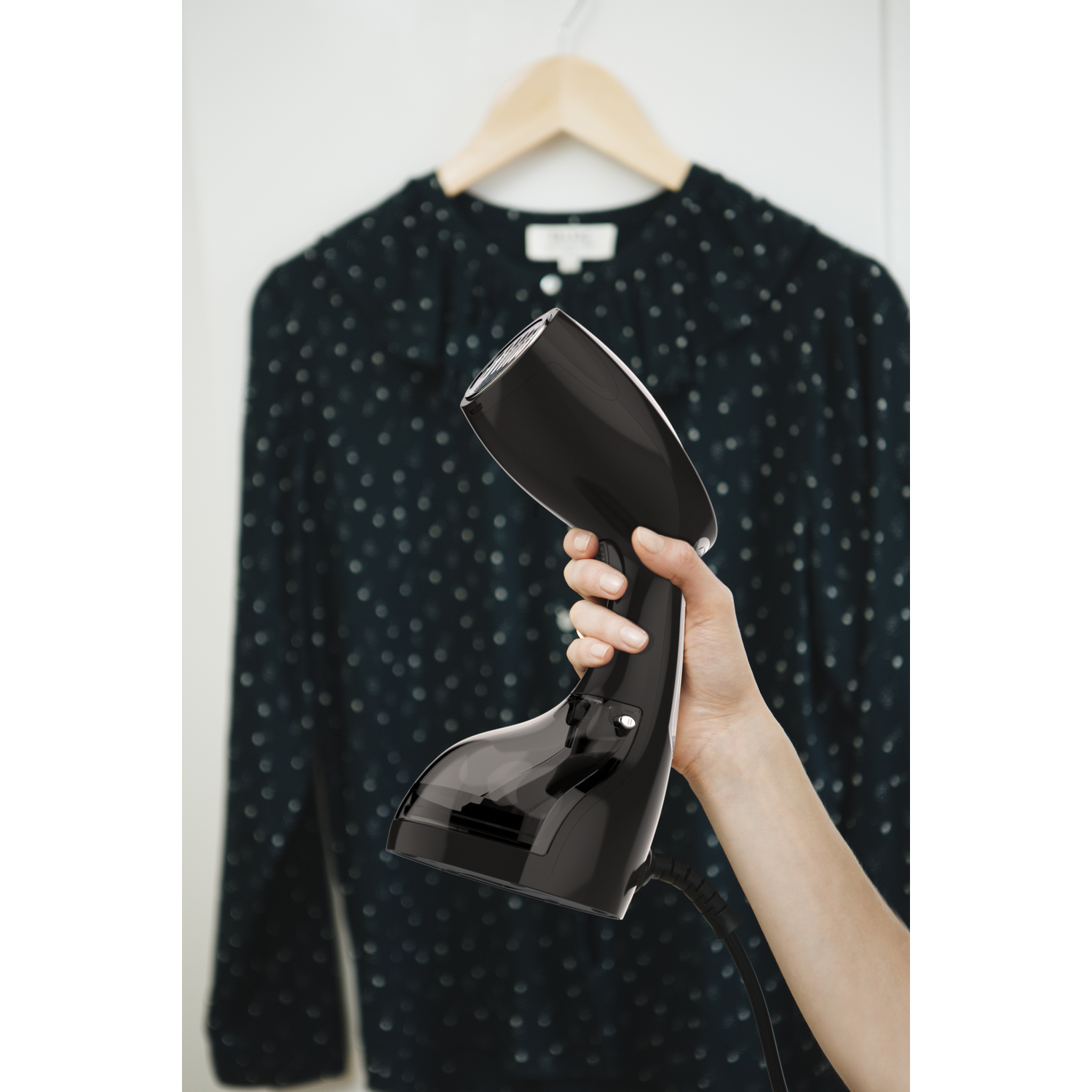 De hand van een persoon houdt een zwarte kledingstomer vast. Een gestippeld overhemd hangt op de achtergrond.