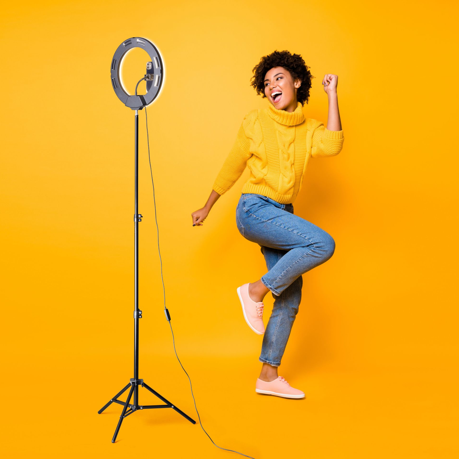 Una mujer baila cerca de una luz de anillo en un soporte, con un suéter amarillo y jeans azules sobre amarillo.