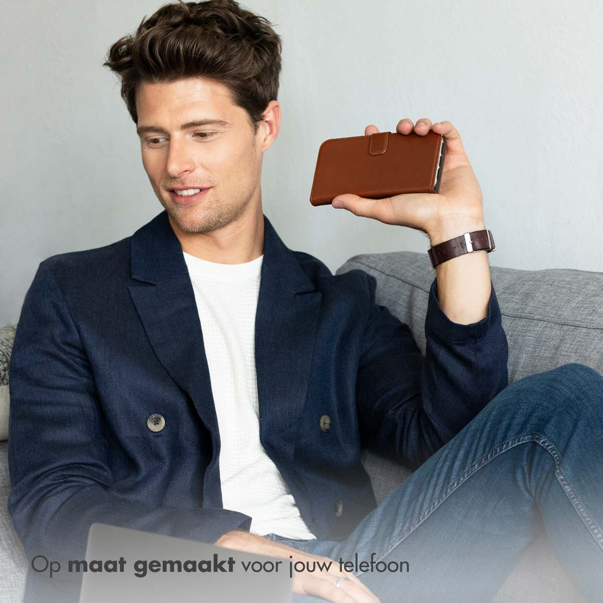 Mann in blauem Blazer und Jeans hält eine braune Handyhülle. Er sitzt auf einem Sofa.