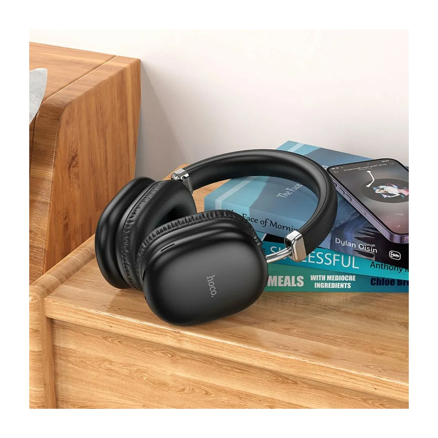 Schwarze kabellose Kopfhörer auf einem Holztisch, neben Büchern und einem Telefon mit Musik-App.