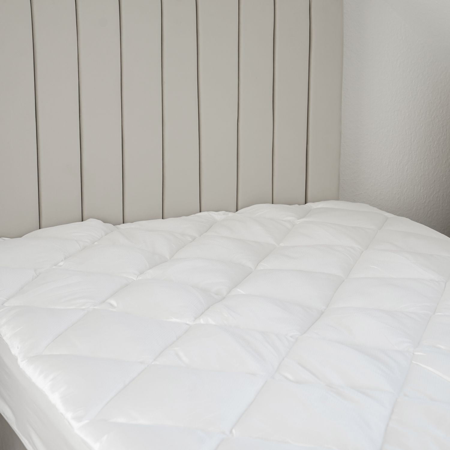 Nahaufnahme eines weißen, gesteppten Matratzenschoners auf einem Bett. Das Kopfteil ist sichtbar.