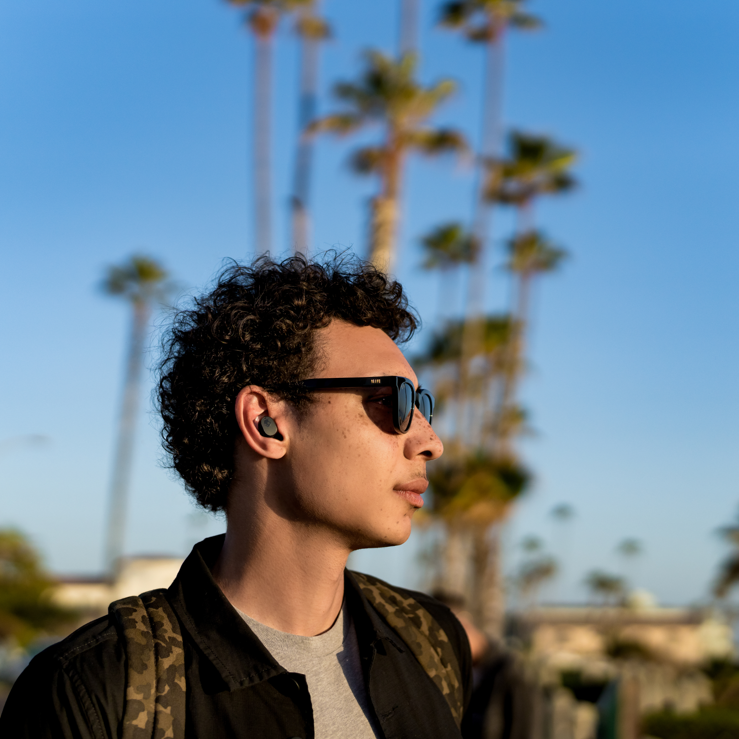 Junger Mann mit Sonnenbrille und Ohrhörern. Palmen im Hintergrund.