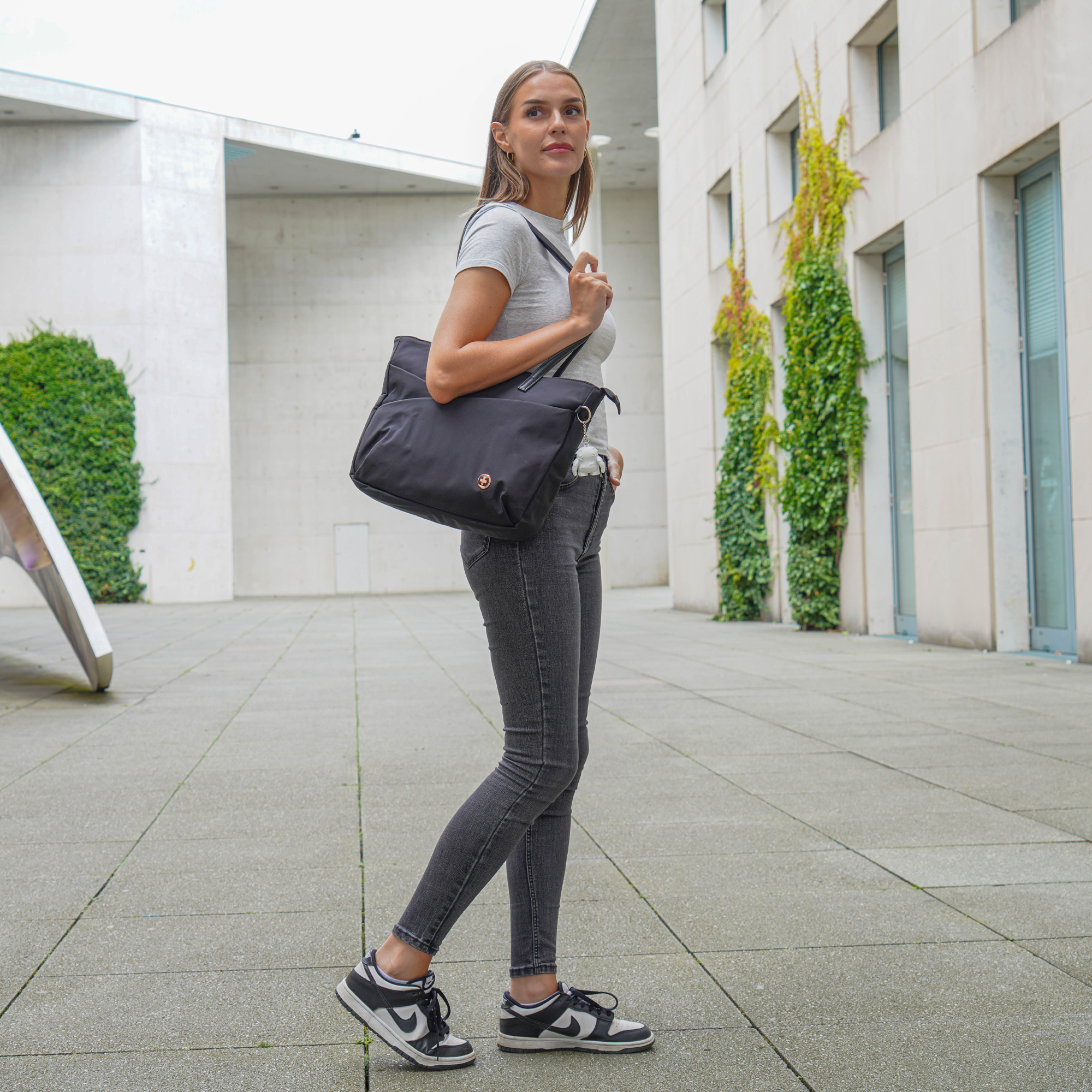 Donna in camicia grigia e jeans porta una borsa nera camminando sul marciapiede, vicino a un edificio moderno.
