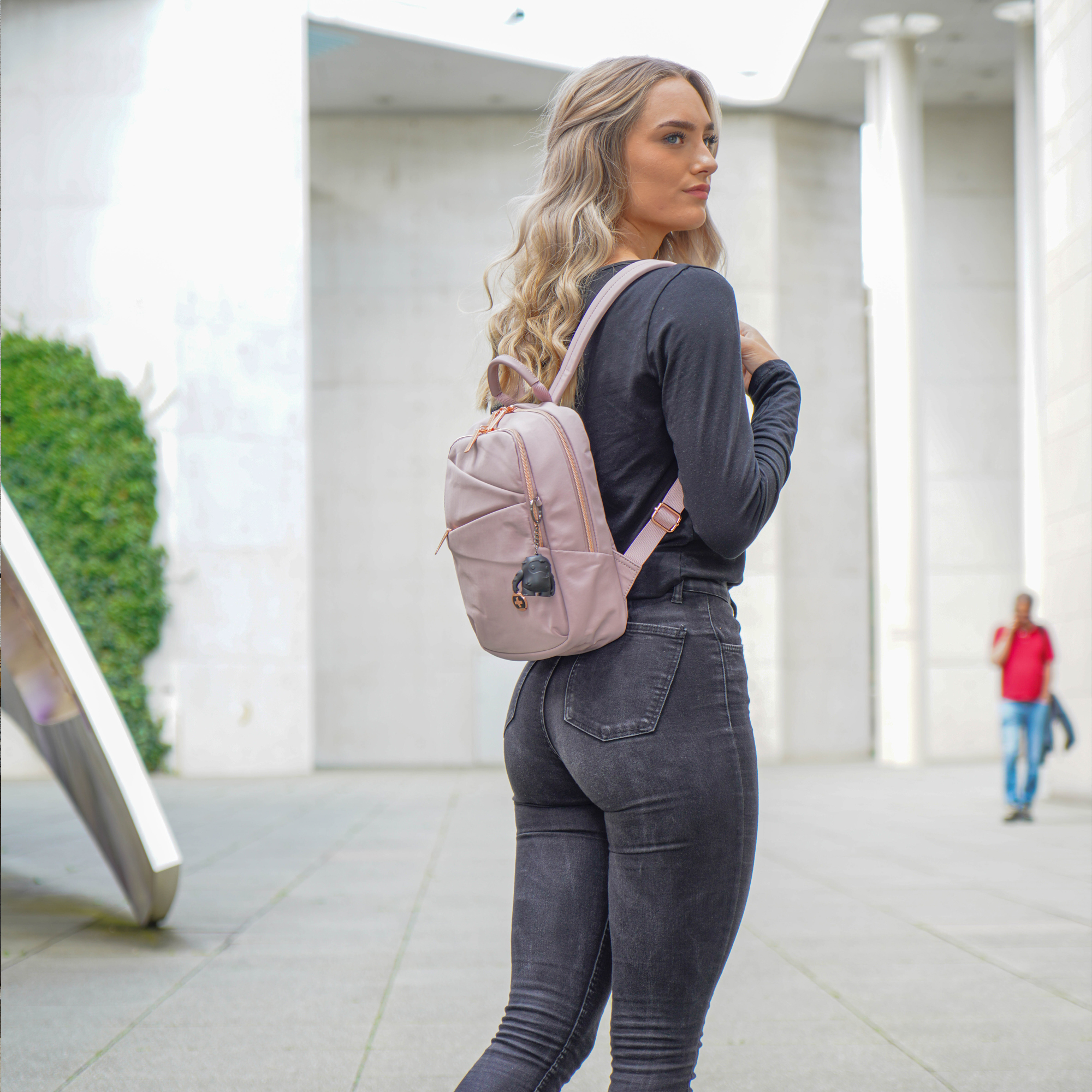 Donna che cammina, con uno zaino rosa e jeans. Sfondo cittadino.