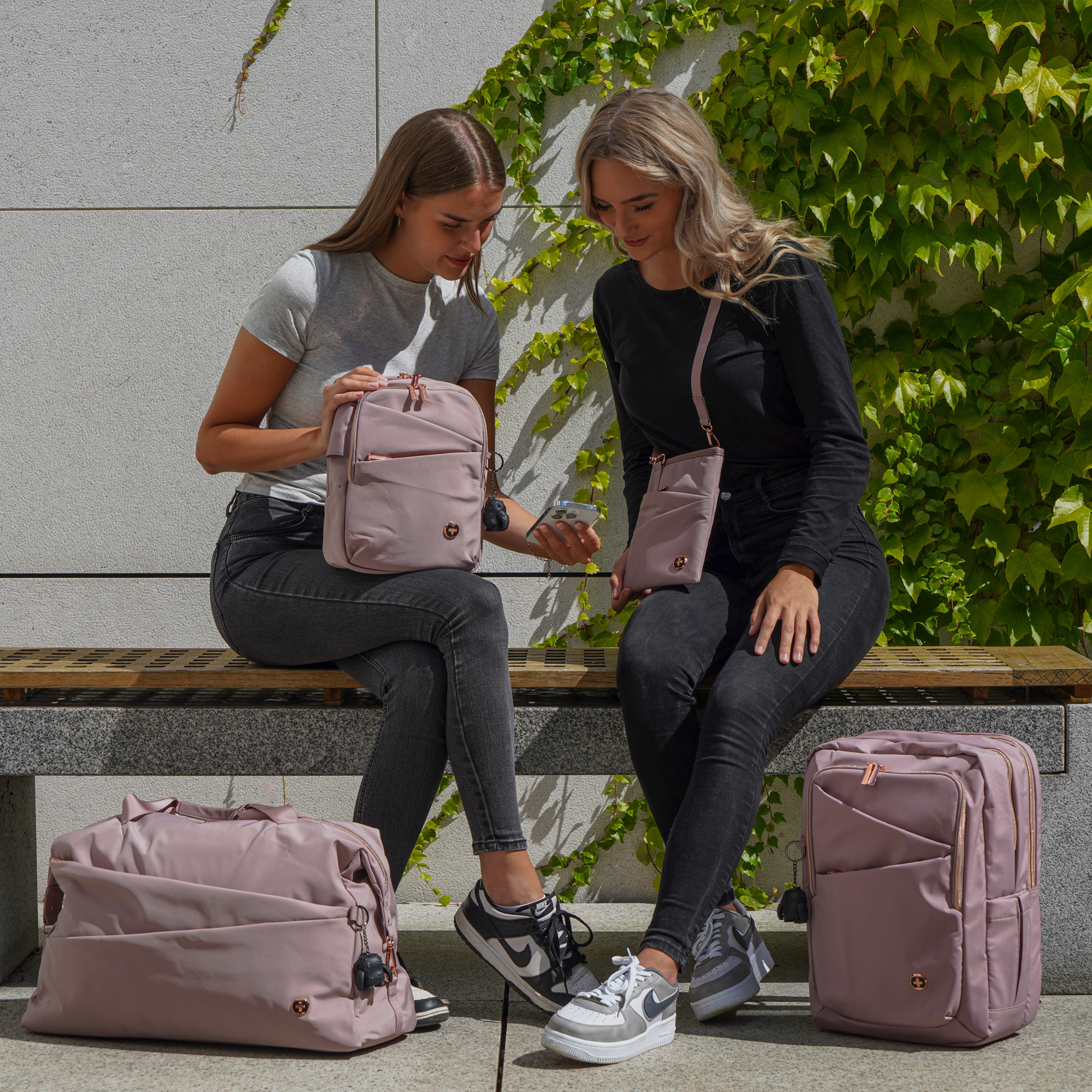 Due donne sedute su una panchina, con bagagli rosa e un muro con piante verdi sullo sfondo.