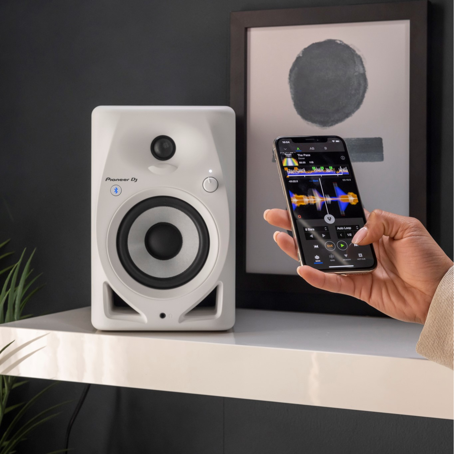 Een witte Pioneer DJ-luidspreker op een plank. Een telefoon toont een muziek-app. Een schilderij hangt erachter.