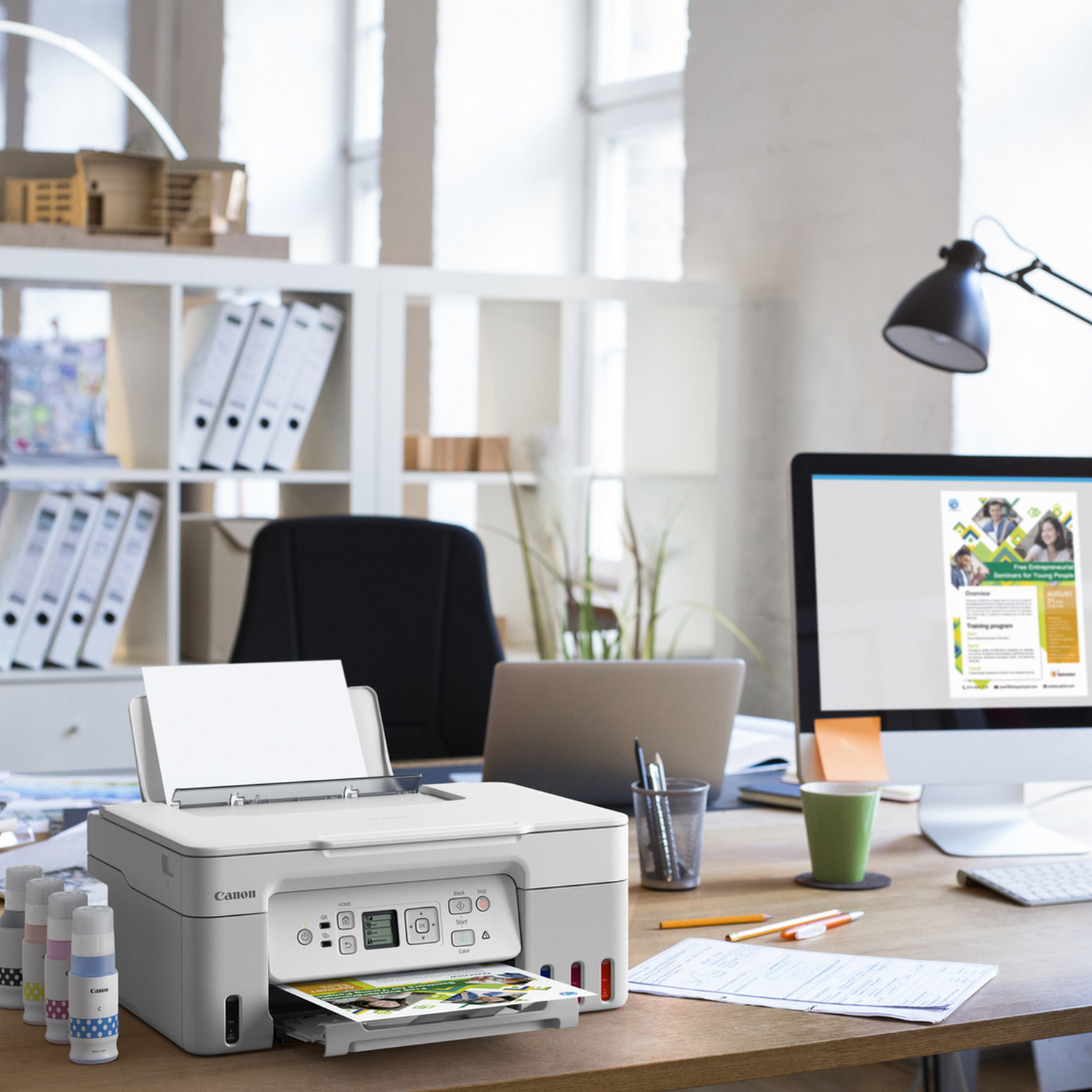 Biała drukarka, wkłady atramentowe i wydrukowany obraz na biurku. Laptop i zielony kubek. Otoczenie biurowe.